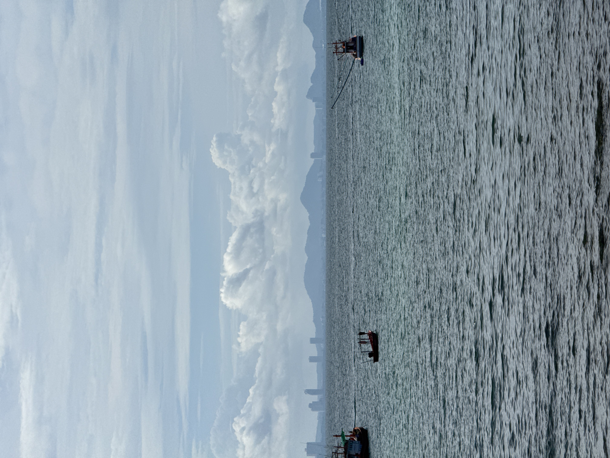 The image shows an ocean view, presumably off the coast of Pattaya City, Thailand. There are several fishing boats on the water. The skyline of the city is faintly visible in the distance, between the boats and the clouds. The sky is cloudy and overcast. The image is rotated 90 degrees clockwise.
