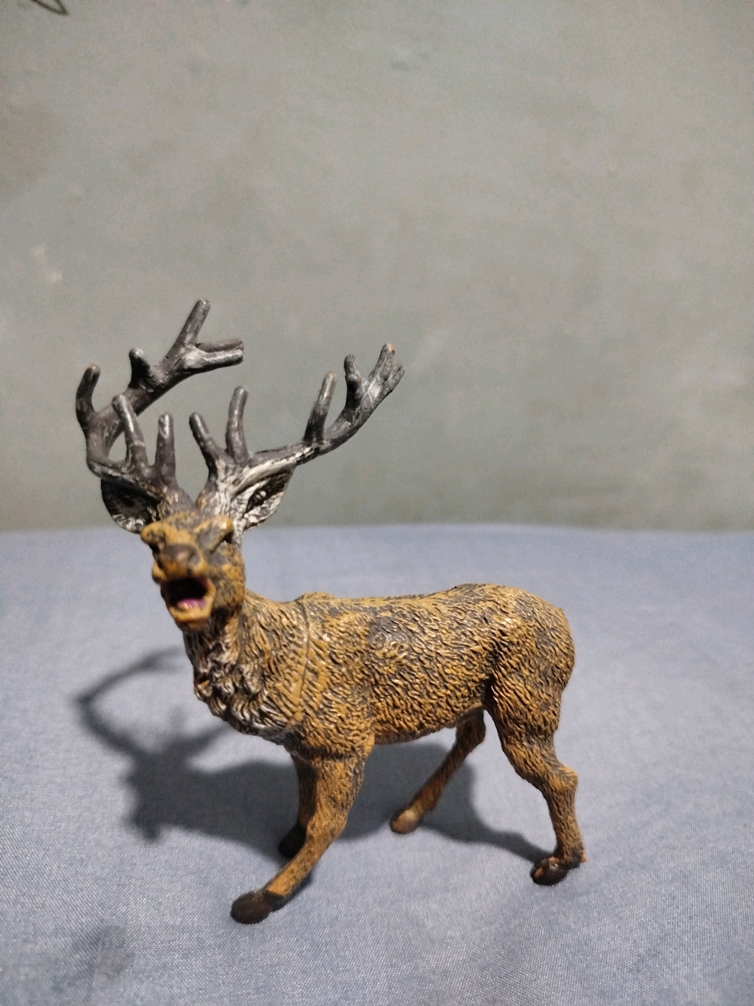 A brown and black toy deer stands on a blue textured surface in front of a grey wall. The deer is positioned to the left of the frame and is facing forward, with its mouth open and tongue visible. It has large antlers and a detailed textured coat. A shadow of the deer is cast on the surface to its left. The background is a plain grey wall, slightly out of focus. The image is taken from a low angle.