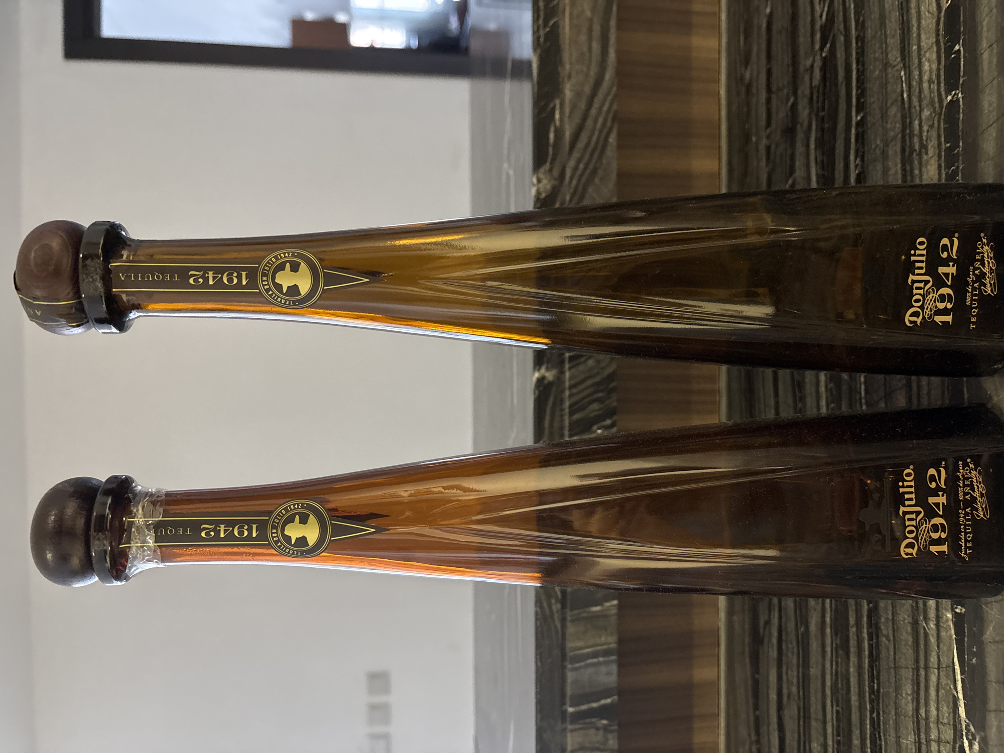 Two bottles of Don Julio 1942 tequila stand upright on a dark, marbled countertop. The bottles are made of amber glass and taper towards the top, each topped with a dark wooden stopper. Both bottles feature a yellow label with "Don Julio 1942" written in prominent gold lettering. The background is a plain, lightly colored wall with two visible light switches. The scene is illuminated by natural light, casting subtle reflections on the glass bottles. The bottles are positioned in a way that suggests they are on display. There are no people present in the image.