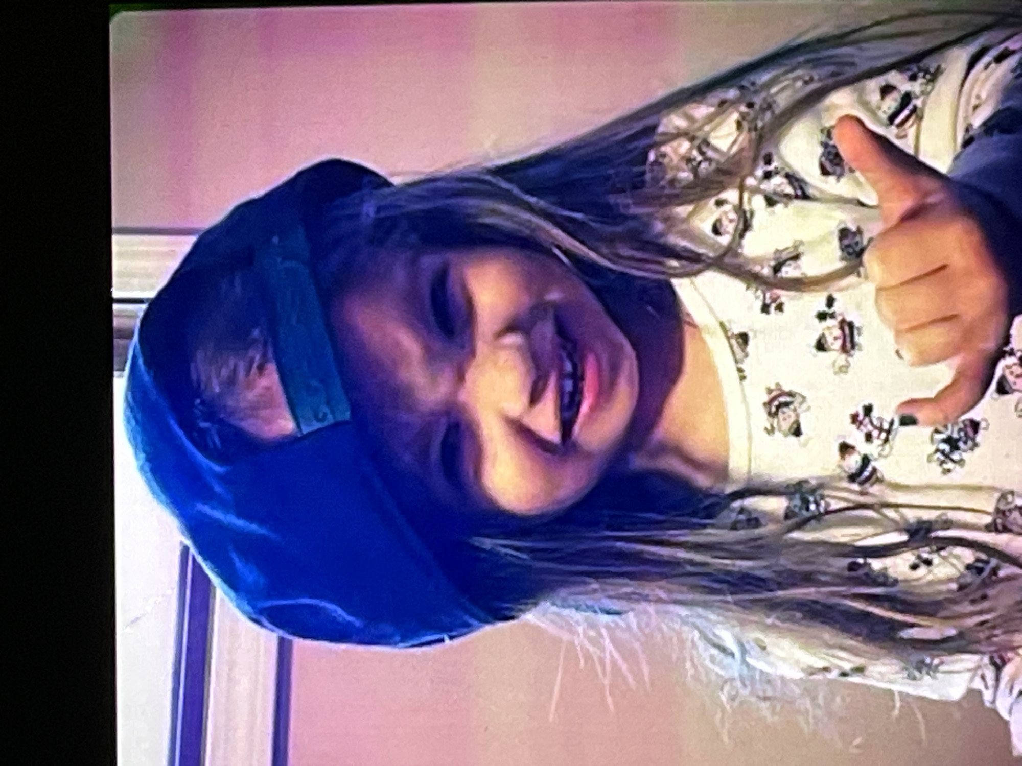 The media file shows a young girl wearing a blue hooded garment and a pajama top with a repeating pattern of cartoon characters. She has long, light brown hair that is spread out around her. Her head is tilted to the side, and her face is partially obscured by shadows and the angle of the image. She appears to be smiling or laughing, with her mouth open. Her right hand is raised, and she is giving a thumbs-up gesture. The background is out of focus and appears to be an indoor setting with soft, warm lighting, suggesting it might be nighttime or evening. The image is a photograph, taken from a slightly elevated angle, and the quality suggests it might be a digital photo from a casual setting. There is no visible text in the image. The overall mood is cheerful and playful, conveyed by the girl's expression and gesture.
