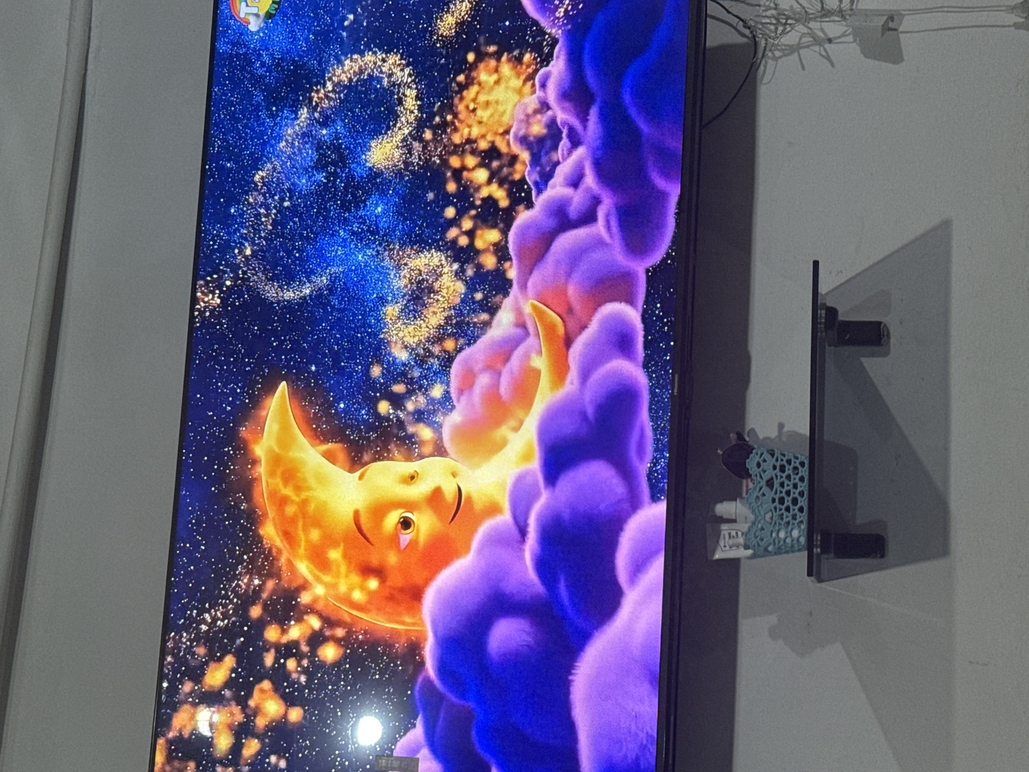 A television screen displays an animated crescent moon character with a smiling face and bright orange skin, set against a dark blue, star-filled night sky. The moon is partially obscured by fluffy, purple and pink clouds. Below the television, a dark gray wall features a small, floating black shelf. On the shelf sits a light blue, woven plastic basket containing several small white bottles and a dark object, possibly a makeup brush. Wires are visible on the wall to the right of the shelf. The scene appears to be an interior view, likely a room, in Chardara, Uzbekistan.