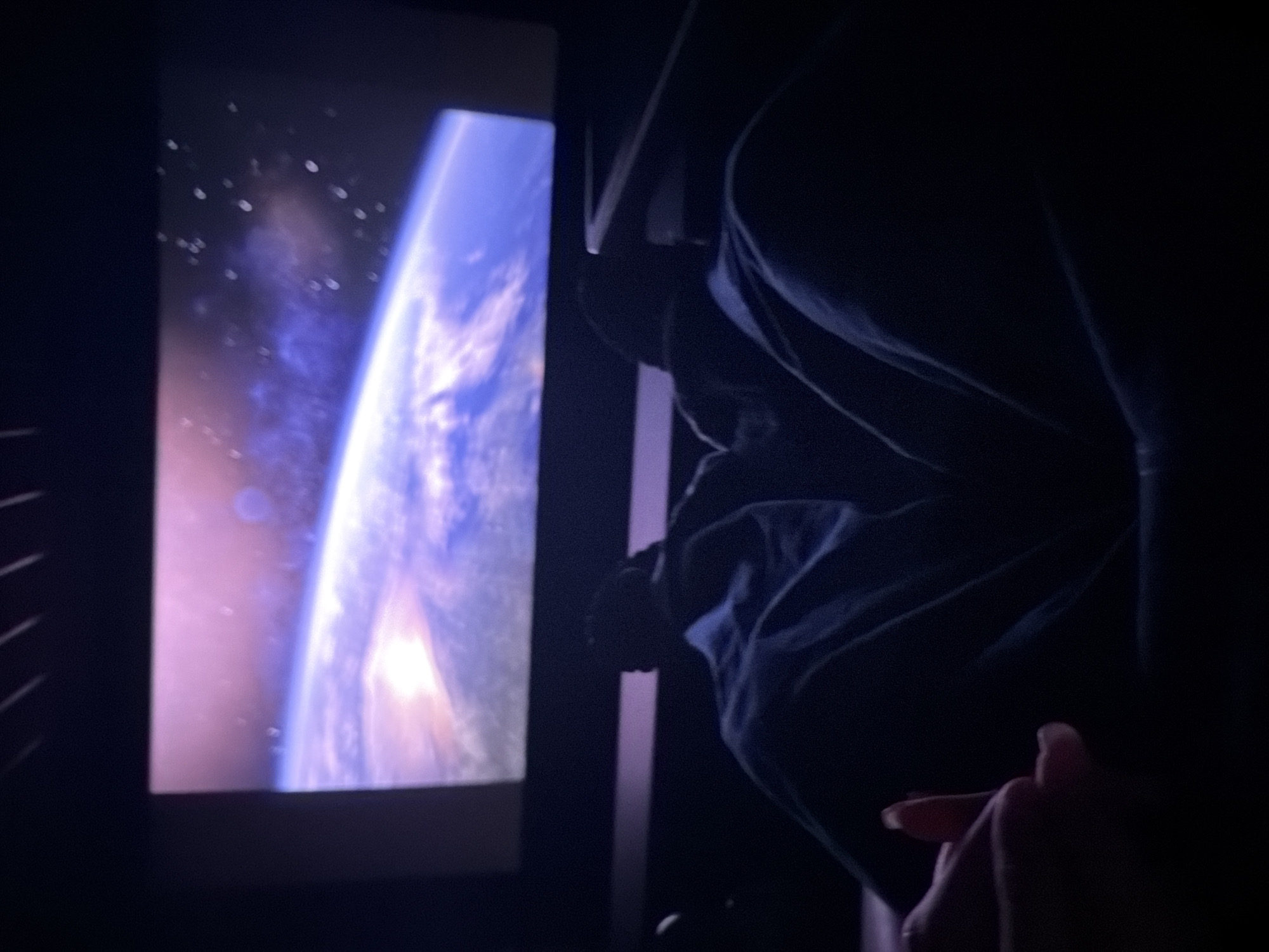 The image shows a partially obscured view of a screen displaying a celestial image of a planet from space, possibly Earth. To the right of the screen, a person's torso and hand are visible in deep shadow. The person's clothes appear to be dark-colored. The surrounding environment is also dark, suggesting an indoor setting with low lighting. Without further information, it is not possible to determine the location in Sofia, Bulgaria.