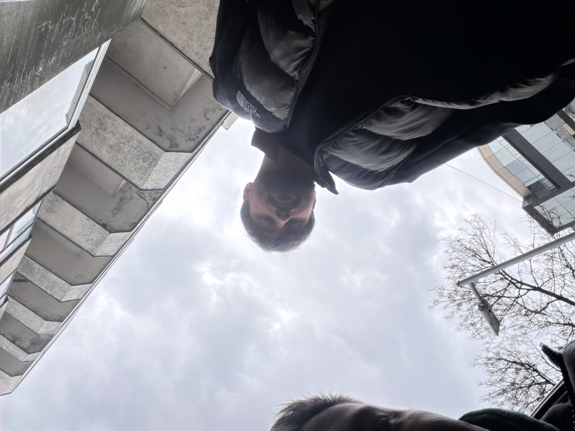 The image, taken from a low angle, depicts a man in a black North Face puffer jacket standing outside in Varna, Bulgaria. The building with protruding concrete balconies is on the left. Another building with a glass facade is visible in the background. Bare tree branches are visible against the cloudy sky. Another person is visible near the bottom of the frame.