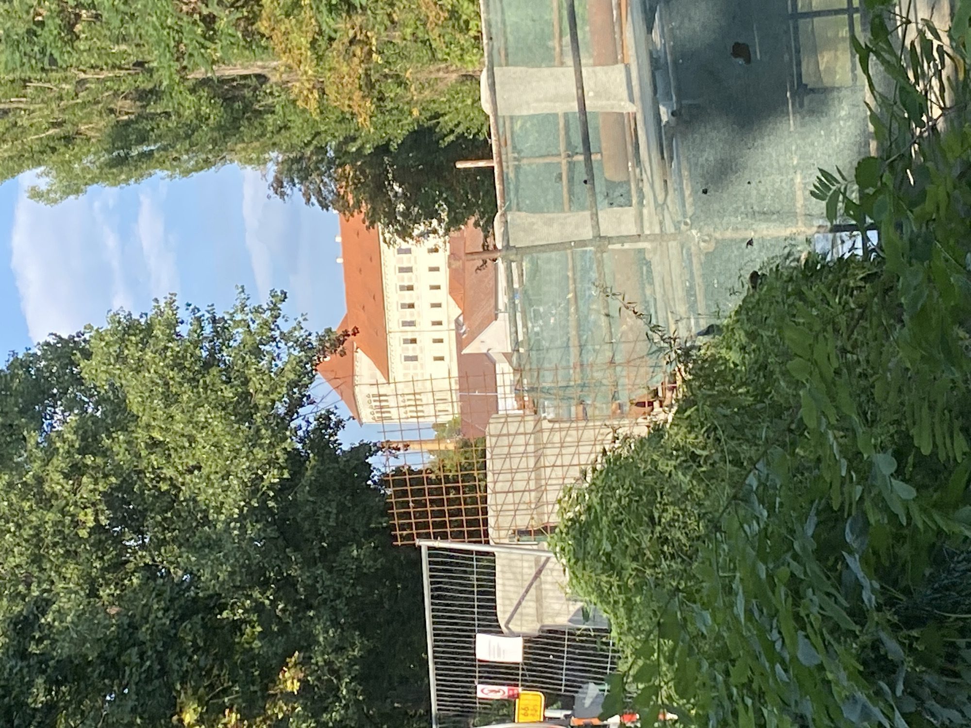 The image shows a construction site in front of a large, light-colored building with a red-tiled roof, partially obscured by trees. The building appears to be a historical structure, possibly a castle or palace. In the foreground, there is a metal fence with a wire mesh, indicating construction or renovation work. Green netting is also visible, stretched over scaffolding. Lush green foliage, including trees and bushes, surrounds the site and partially obstructs the view of the building. The sky is partly cloudy and blue. The scene is likely located in or near a park or garden area due to the abundant greenery.