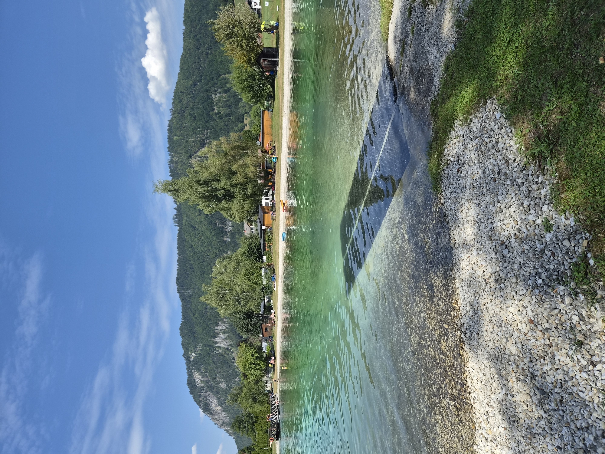 This image depicts a serene lakefront scene on a sunny day in Landl, Austria. The foreground is dominated by a clear, shallow body of water with a pebble-strewn bottom, gradually deepening into a vibrant turquoise hue. A ramp, likely for watercraft or accessibility, extends from the shore into the water, casting a distinct shadow.
The middle ground features a sandy beach area with scattered sunbathers and a few small structures, possibly cabins or changing rooms. Beyond the beach, a line of trees creates a lush green backdrop, with a few taller trees standing out. In the distance, a majestic, tree-covered mountain range rises against a bright blue sky dotted with wispy white clouds. The overall atmosphere is one of a peaceful recreational area.