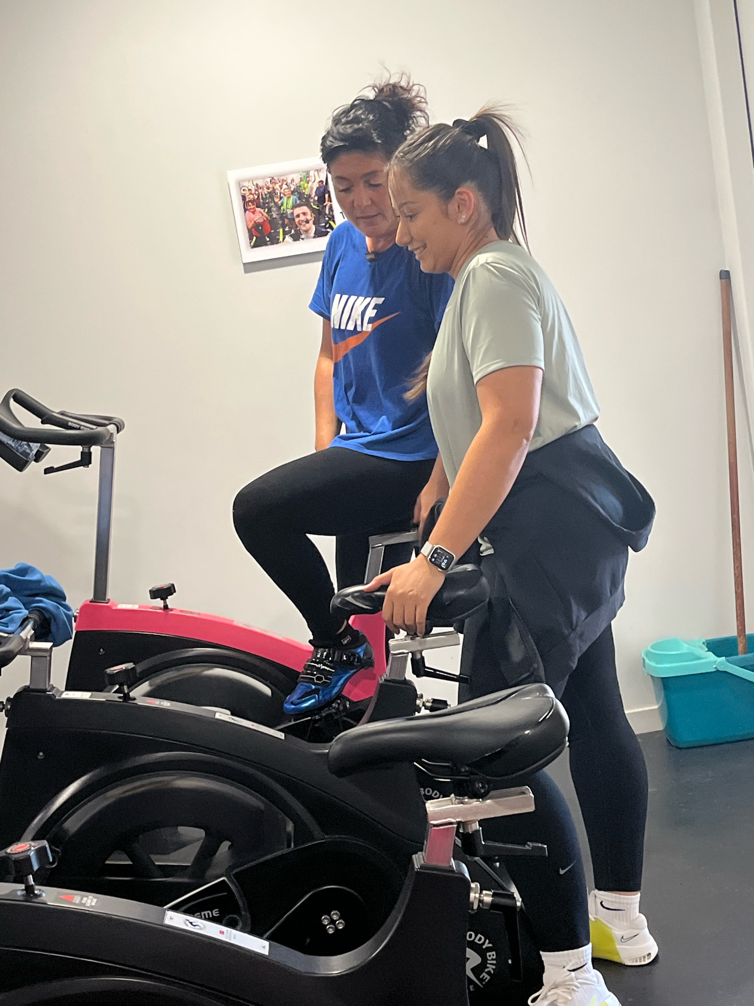 Two women are exercising on stationary bicycles. The woman on the left is wearing a blue t-shirt with the Nike logo and black leggings. The woman on the right is wearing a light green t-shirt, black leggings, and white socks with a yellow heel. She has a smartwatch on her left wrist. They appear to be in a gym or fitness studio. In the background, there is a framed picture on the wall of a group of people, and a blue bucket with cleaning supplies. The location is Saint-Galmier, France.