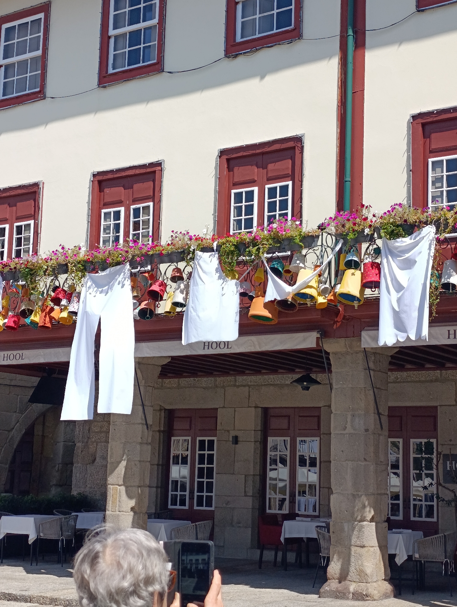 A wide shot captures the exterior of a building in Guimarães, Portugal, under a bright sky. The building features a light-colored facade with several windows framed in dark red. Some windows have white panes, while others are dark. A balcony on the second floor is adorned with an abundance of colorful, bell-shaped decorations and two large white pieces of fabric hanging down. Below this, on the first floor, is a covered walkway with stone columns and more dark red framed windows. Tables with white tablecloths and chairs are set up in the area beneath the walkway. In the foreground, the back of an older person's head and shoulders are visible as they hold up a smartphone, appearing to take a picture of the scene. The overall impression is a lively and festive atmosphere, possibly at an outdoor dining area or marketplace.