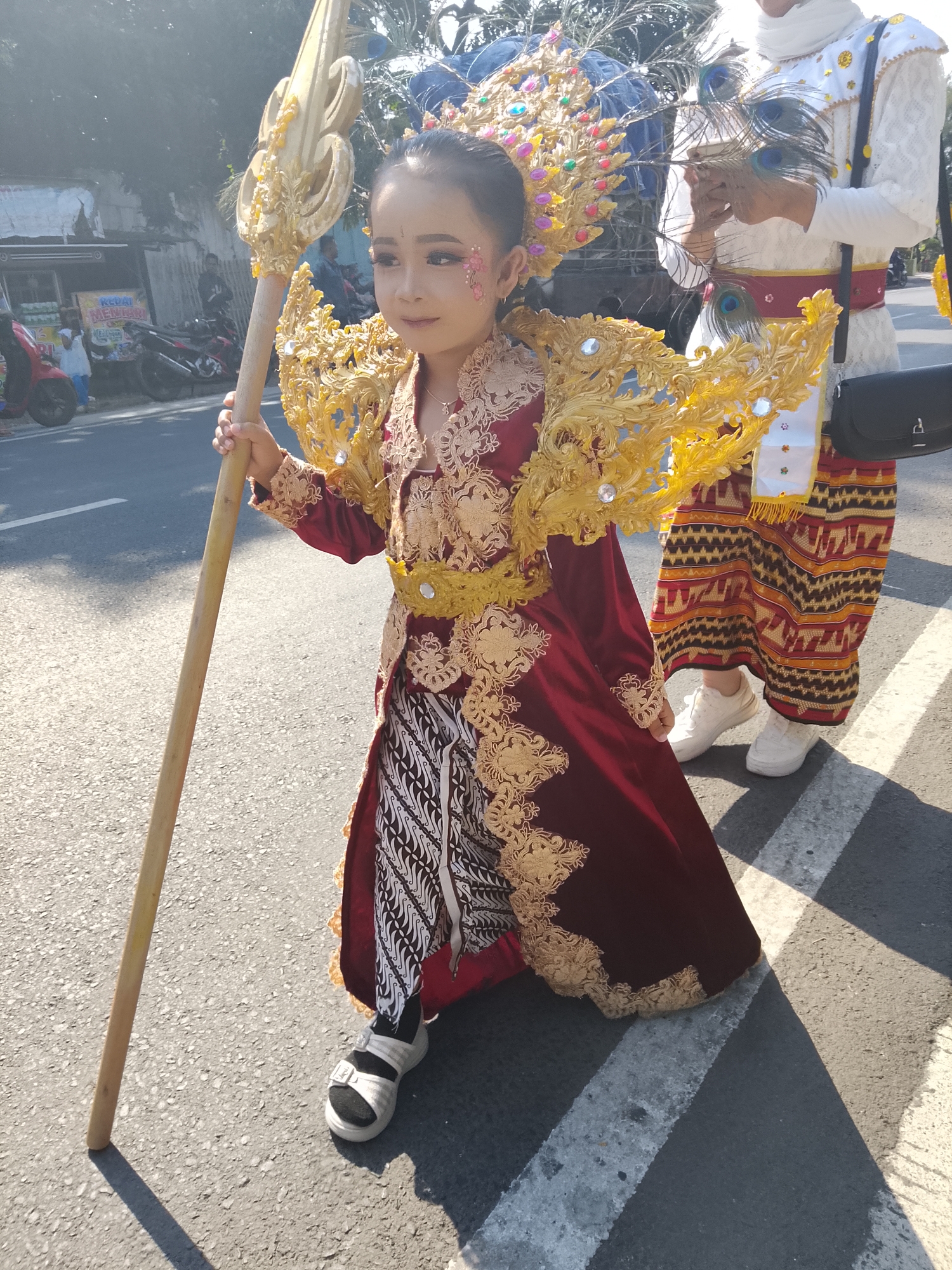 A young girl dressed in a red and gold ceremonial costume, featuring an elaborate headdress, stands on a street in Buduan, Indonesia. She holds a tall, golden staff. Behind her, another person in white ceremonial attire with a red sash and patterned fabric skirt, is also visible. The scene appears to be a parade or cultural event, with a paved road, white markings, and trees lining the background. Sunlight casts shadows on the ground.