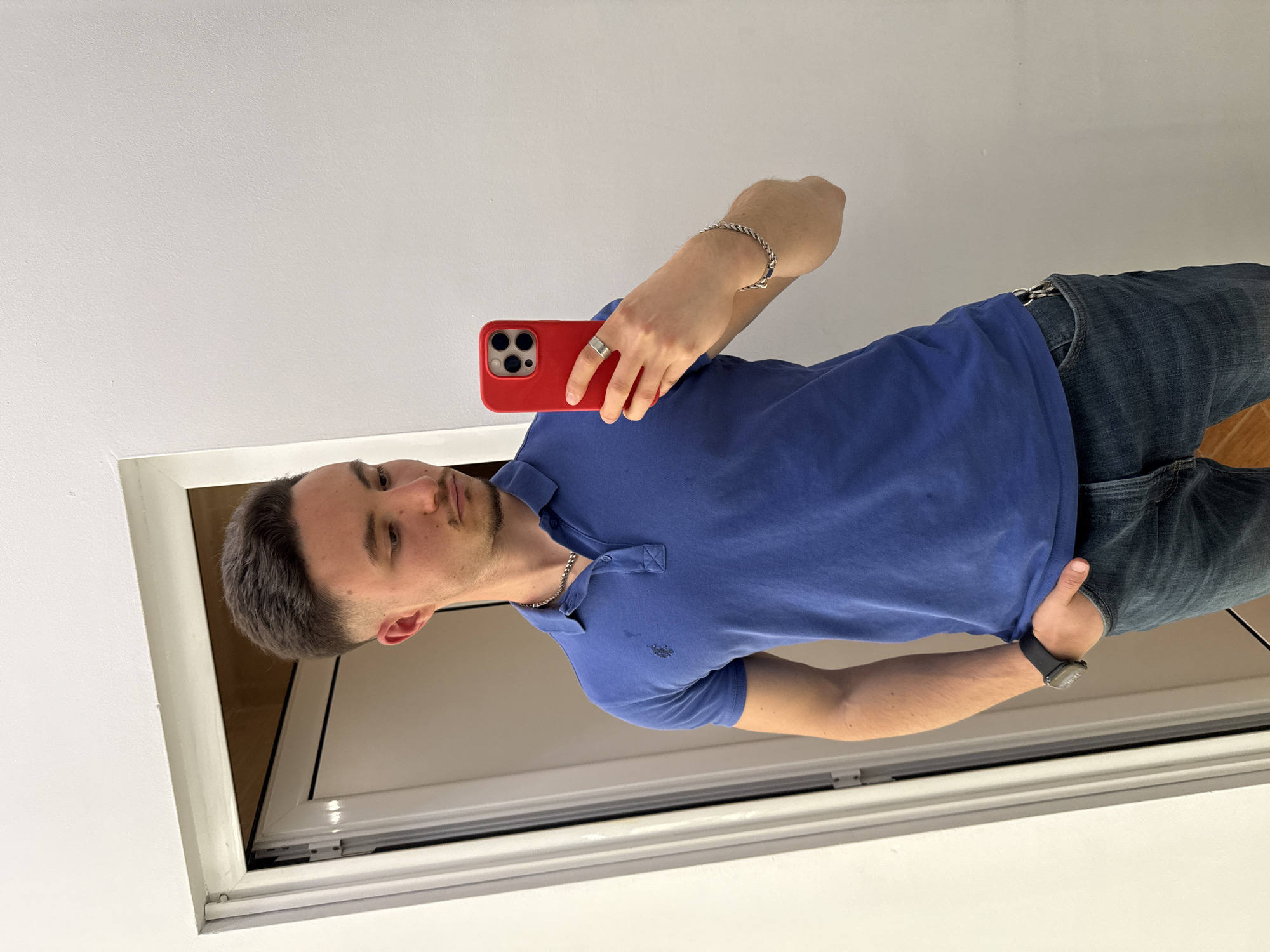 A young man is depicted standing indoors, likely captured in a selfie taken in a mirror or reflective surface. He has short, dark hair and short facial hair. He is wearing a short-sleeved, royal blue polo shirt with a subtle, dark emblem on the left chest, paired with blue denim jeans.
He is accessorized with a thin silver chain around his neck. On his left hand, he wears a silver ring on his ring finger and holds a red-cased smartphone (featuring a triple-lens camera system), angled upwards towards himself. On his right wrist, he wears a silver chain bracelet and a black smartwatch, with his right hand resting in his jeans pocket.
The background consists of a plain white wall, and a white window or door frame with a dark inner edge, suggesting a contemporary interior space. A small section of a warm-toned wooden floor is visible in the lower right corner. The lighting appears bright and even, indicating daytime. The man looks directly into the camera with a neutral expression. No distinct text is visible on his clothing or accessories, apart from the indistinct logo on his shirt. The image provides no intrinsic geographical cues, but is contextualized as being taken in Sofia, Bulgaria.