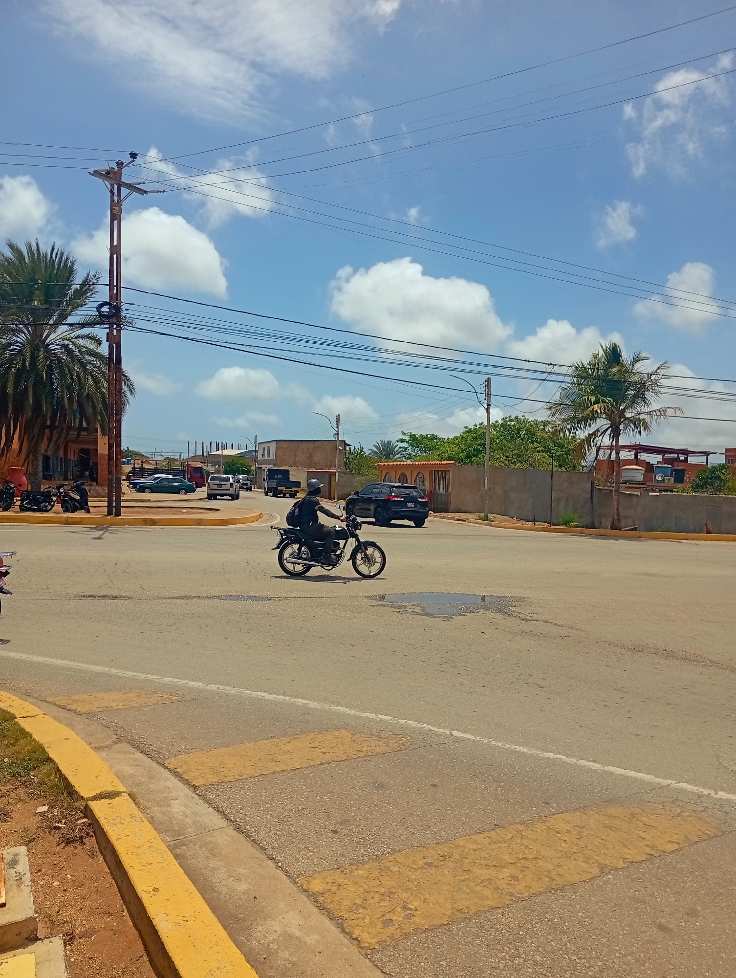This is a medium-shot, eye-level, vertical photograph taken in Juan Griego, Venezuela. The scene depicts an intersection on a sunny day with a clear blue sky dotted with white clouds. A person is riding a motorcycle in the foreground, moving from left to right across the frame. The rider is wearing a black helmet and a backpack. The motorcycle is black and has spoke wheels. The road surface is asphalt with a marked pedestrian crossing featuring yellow stripes. A curb with yellow paint runs along the left side of the road. In the background, various vehicles are visible, including a black SUV, a white SUV, a red truck, and several parked motorcycles in front of a building with palm trees. Power lines are strung across the upper portion of the image, supported by a wooden utility pole on the left. Buildings with windows and rooftops are visible in the middle and far background, suggesting an urban or semi-urban environment.