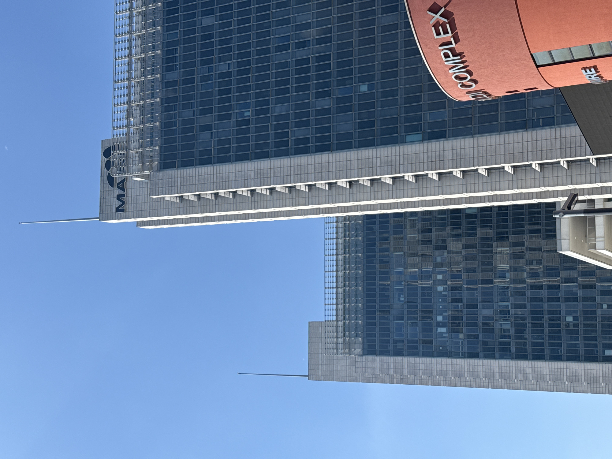 The image displays a modern architectural scene under a clear, bright blue sky, rotated ninety degrees to the left. The primary focus is on two contemporary skyscrapers. The foreground building features a dark, grid-patterned glass facade that reflects parts of another building. Below the glass section, there's a multi-tiered concrete or stone structure with small, repetitive protrusions. A slender, metallic spire extends upwards from a lower section of this building.
On the main building, near the spire, the letters "MA" are visible in a dark font, accompanied by a logo resembling three interconnected circles. To the right, a prominent, curved, terracotta-colored structure bears white, three-dimensional lettering that clearly spells "DI COMPLEX". This red structure appears to be an integral part of or attached to one of the buildings.
The scene captures a sunny daytime, indicated by the vibrant blue sky and crisp lighting on the building surfaces. There are no people or explicit activities depicted. The setting is urban, with the architecture suggesting a modern city. The overall impression is one of sleek design and towering structures, characteristic of a business district. The location is Milan, Italy.