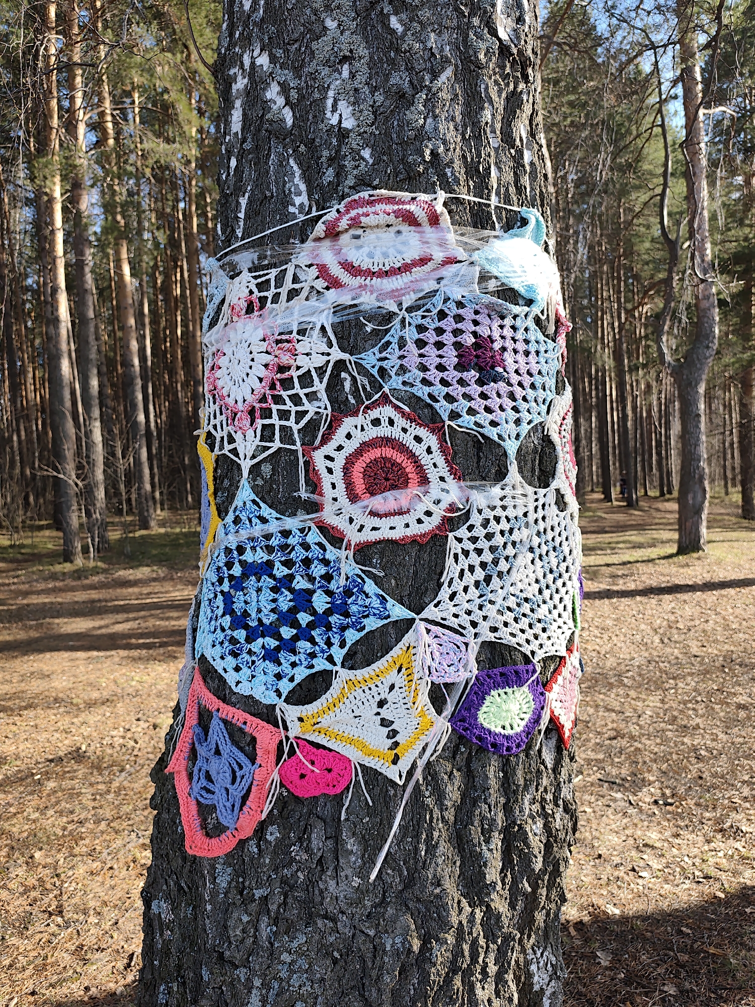 A tree trunk is wrapped in a colorful crocheted textile installation in a wooded area, likely in Chaikovsky, Russia. The tree's bark is visible beneath the fabric. The textile appears to be composed of many crocheted granny squares and circular patterns of various colors, including white, blue, pink, red, purple, yellow, and green. A transparent material is used to secure the textile to the trunk. The background consists of other trees and leaf-covered ground, suggesting a forest or park setting. The lighting indicates a sunny day.