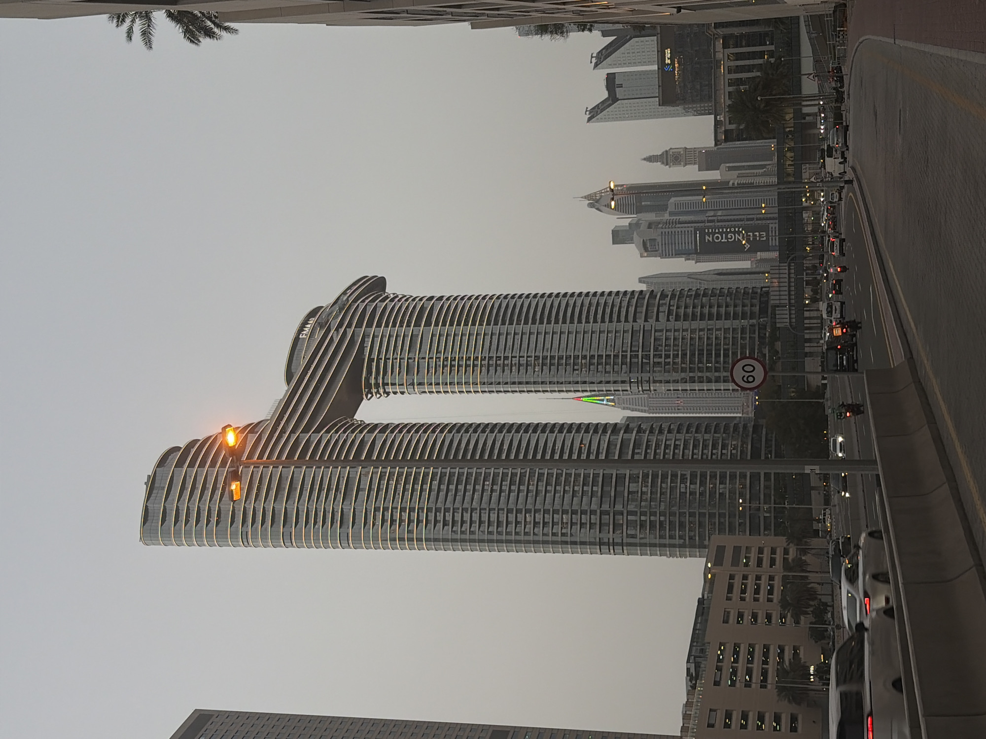 The image shows a partial cityscape of Dubai, United Arab Emirates. The most prominent feature is a modern, interconnected skyscraper with two towers joined at the top by a horizontal structure, forming a distinctive "H" shape. The towers have a facade of vertical stripes. The sky is overcast, casting a muted light on the buildings. A street runs along the right side of the frame with traffic moving along it. Other buildings are visible in the background. A 60 sign is on the side of the road.
