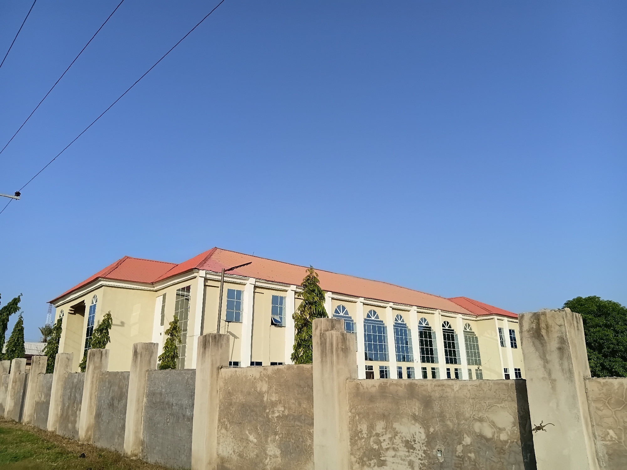 The image depicts a building with a red roof, light yellow walls, and large windows. A concrete fence with vertical pillars runs across the foreground. Electrical wires are visible in the upper left corner against a clear blue sky. Trees are planted along the side of the building, and some are visible on the left and right edges of the frame. The scene appears to be set in Jalingo, Nigeria, characterized by the architecture and vegetation.