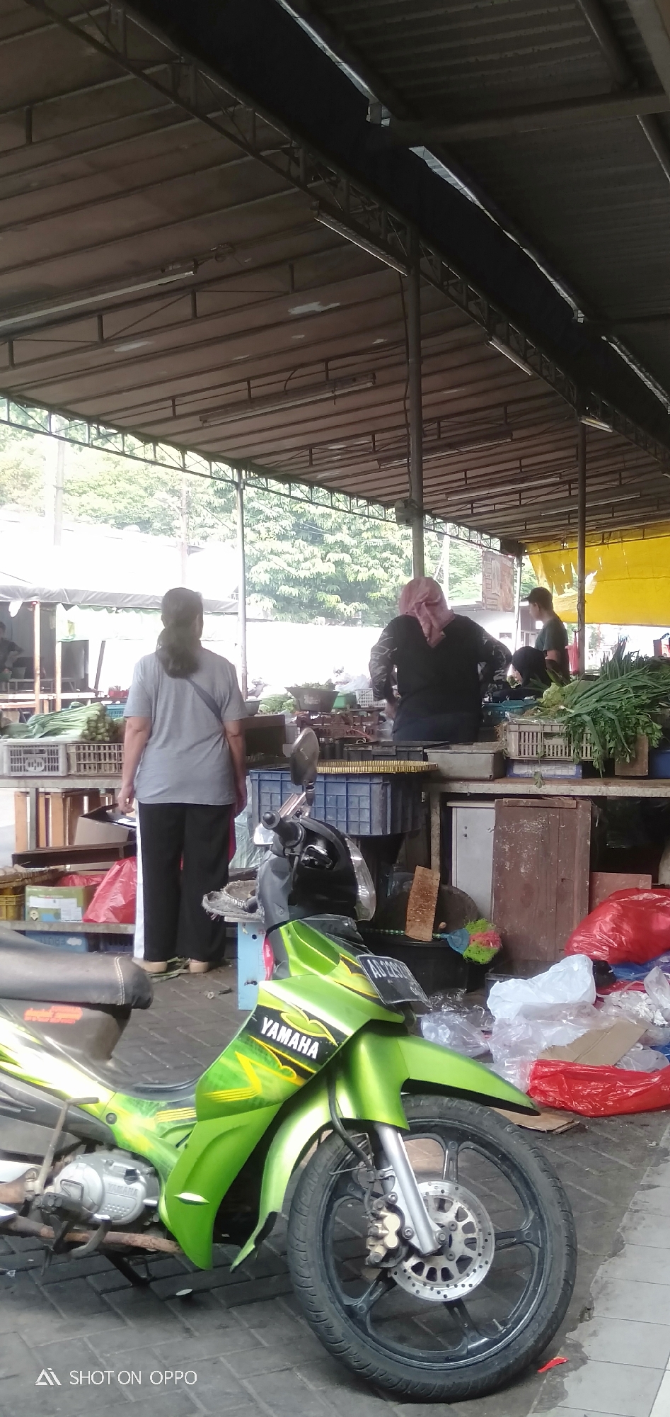 The image shows an outdoor market scene. In the foreground, a bright green Yamaha motorcycle is parked. The motorcycle has the "YAMAHA" logo visible on its side. Behind the motorcycle, several people are visible, appearing to be vendors and shoppers. One person in a grey shirt and black pants stands facing away from the camera, looking towards the market stalls. Further back, two individuals are behind a counter, one wearing a pink headscarf and the other a dark shirt. They appear to be tending to goods displayed on tables. The market is covered by a corrugated metal roof supported by metal beams. Various produce, such as leafy greens and what appear to be bananas, are arranged on tables and in crates. Plastic bags, some red and some white, are scattered on the ground. The ground is paved with bricks. The background reveals trees and foliage, suggesting an urban environment with greenery. The presence of the motorcycle and the open-air market structure suggest a typical scene in a city in South Jakarta, Indonesia.