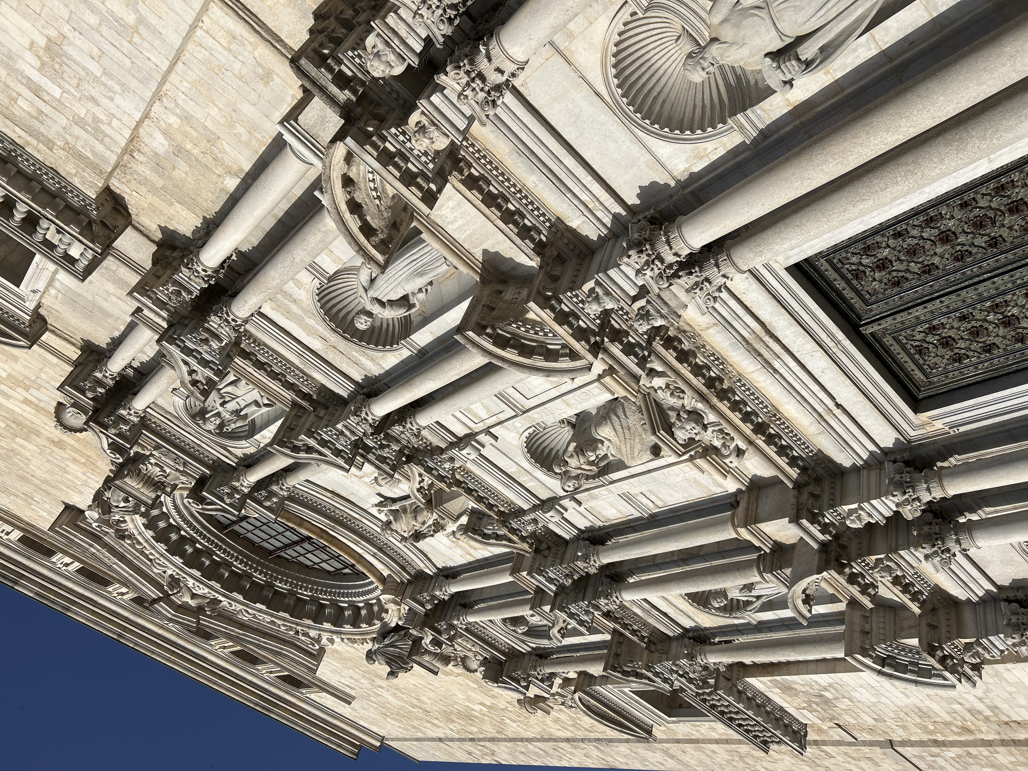The image shows a low-angle, close-up view of the facade of the Girona Cathedral in Girona, Spain. The facade is constructed of light-colored stone with intricate Baroque architectural details. Numerous columns, pilasters, niches, and sculptures adorn the building. Several statues of human figures are visible within the niches, and decorative elements such as cherubs and ornate carvings are present. A large, ornate double door is visible at the bottom right of the frame, featuring decorative metalwork. The sky above is a clear, deep blue. The lighting suggests it is daytime, with strong shadows indicating the sun's position. The perspective is tilted, giving a dynamic and dramatic impression of the cathedral's grandeur.