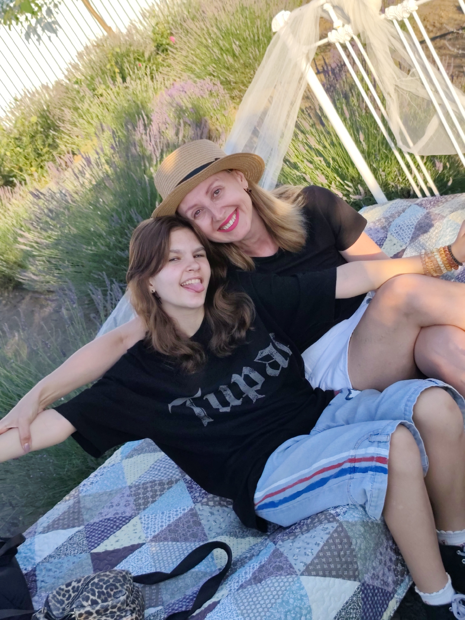 In an outdoor scene bathed in warm, late afternoon light, two females are seated on a colorful patchwork quilt in a field of lavender. The woman in the foreground, on the left, is a teenager with medium-length brown hair and is sticking her tongue out, looking at the camera with a playful expression. She is wearing a black t-shirt with the word "Tupac" printed in large, gothic-style letters across the chest, and light blue shorts with red and blue stripes. The woman behind her, on the right, has blonde hair and is wearing a straw hat and a black t-shirt. She is smiling brightly and has her arm around the teenager. She is wearing several bracelets on her wrist.
Behind the subjects, a white metal structure resembling a bed frame is draped with sheer white fabric, creating a whimsical canopy. The surrounding area is filled with blooming lavender plants, suggesting a tranquil and scenic location. The overall mood is relaxed and joyful, with the bright sunlight casting a warm glow on the scene. The time of day appears to be late afternoon or early evening, indicated by the quality of light. The location is identified as Nechaiane, Ukraine.