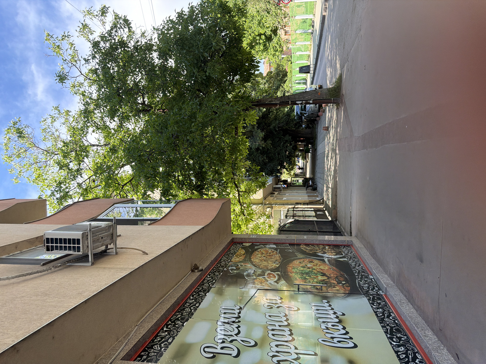 The image depicts a street scene in Pazardzhik, Bulgaria, likely near a commercial establishment. In the foreground, a sidewalk runs alongside a building with a large window display featuring an advertisement for food, with the Cyrillic text "Вземи храна за вкъщи" (Take food home). The building also has an outdoor air conditioning unit mounted on its exterior wall. A sidewalk is visible to the right of the building, shaded by a large tree. Beyond the sidewalk, a street is present with parked cars and a bright green fence lining the opposite side. The sky is visible above, with a few clouds.