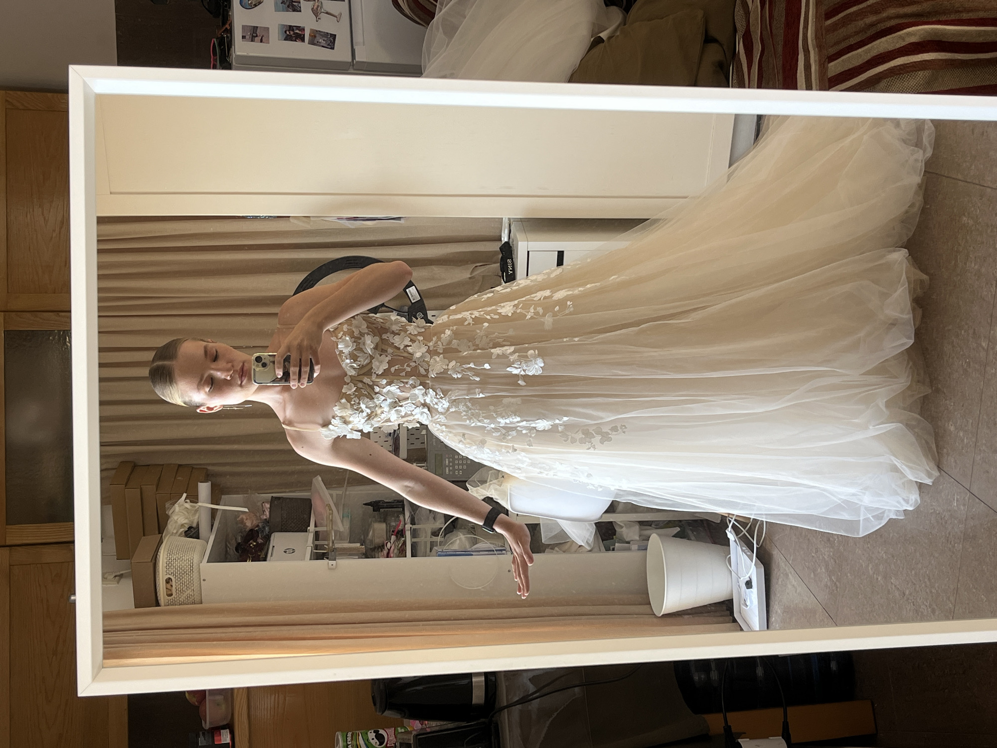 A young woman is captured in a full-body mirror selfie, showcasing a champagne-colored, floor-length wedding dress adorned with delicate white floral appliques. The dress features spaghetti straps and a fitted bodice that flares out into a voluminous tulle skirt. She is holding a smartphone in her right hand, angled to take the photo. Her left hand is extended outwards, touching the side of the mirror. The background is an interior space, likely a dressing room or bedroom. Visible elements include shelving units filled with various items, a white trash can, curtains, a white cabinet, and a refrigerator with magnets on it. The flooring appears to be tiled. The lighting suggests it is daytime, with natural light filtering in from the left and potentially artificial light from a ring light visible to the left of the woman. There are no other people present in the image. The location is identified as Budva, Montenegro.