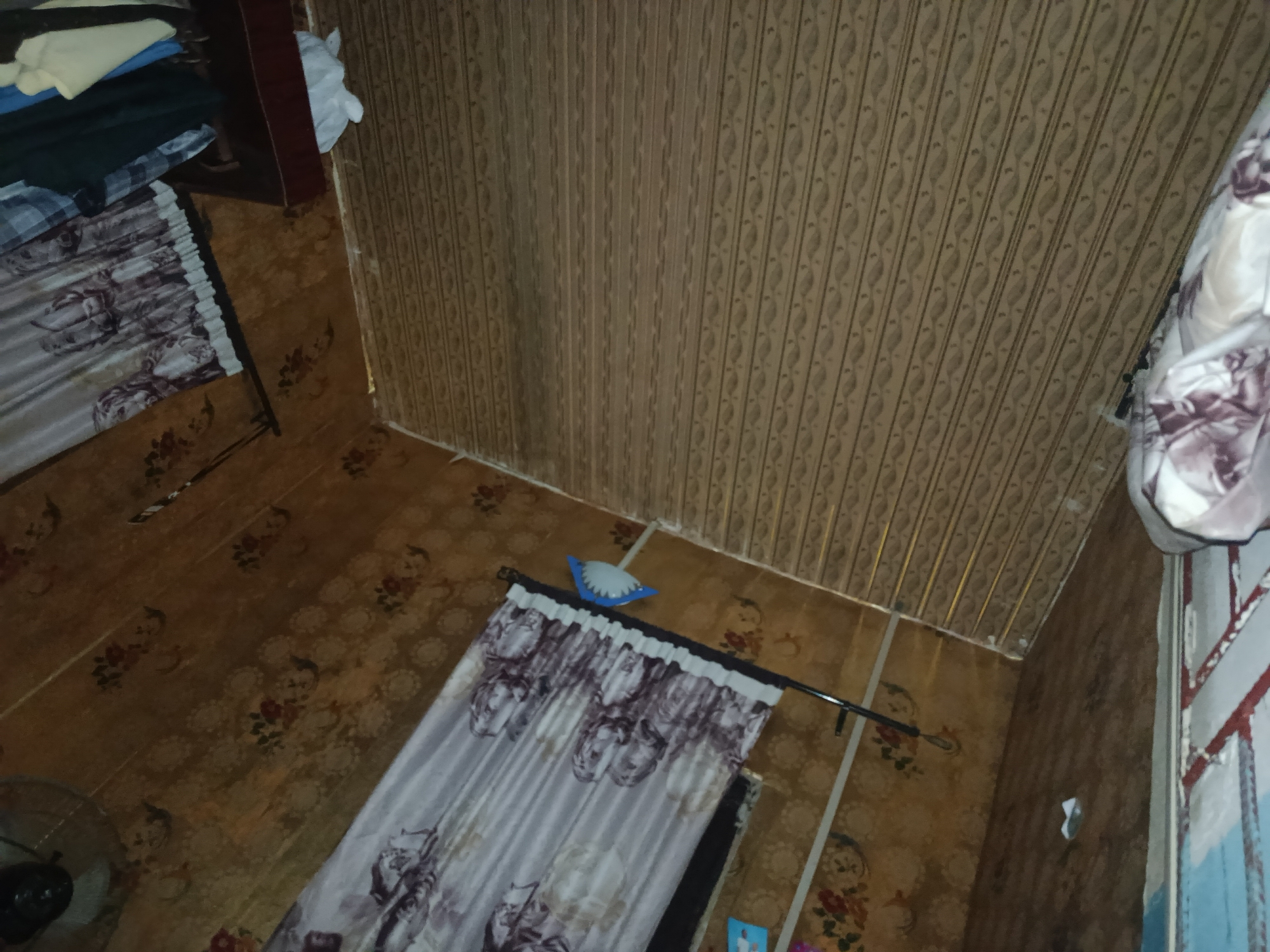 The image shows an interior view of a room, likely a bedroom, located in Afaha Ukwa, Nigeria. The walls and ceiling are covered in patterned paneling. The floor is covered with a floral-patterned linoleum. In the foreground, a bed with patterned curtains is visible. To the left, a stack of clothes is placed on a shelf or piece of furniture. A fan is partially visible in the bottom left corner. A light fixture is attached to the wall near the bed. The scene appears to be a modest living space.