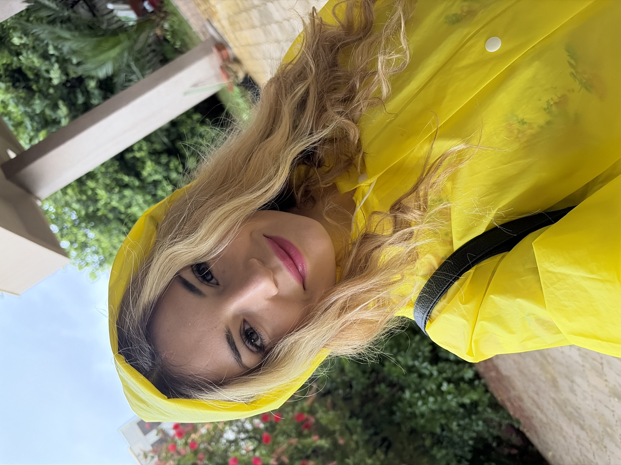 A woman with blonde wavy hair, wearing a bright yellow raincoat with the hood up, looks at the camera. She has pink lipstick. A black purse or bag strap is visible over her right shoulder. In the background is greenery including trees, bushes, and red flowers, a white structure, and a cloudy sky. Given the presence of lush vegetation and building style, this could be in Budva, Montenegro.