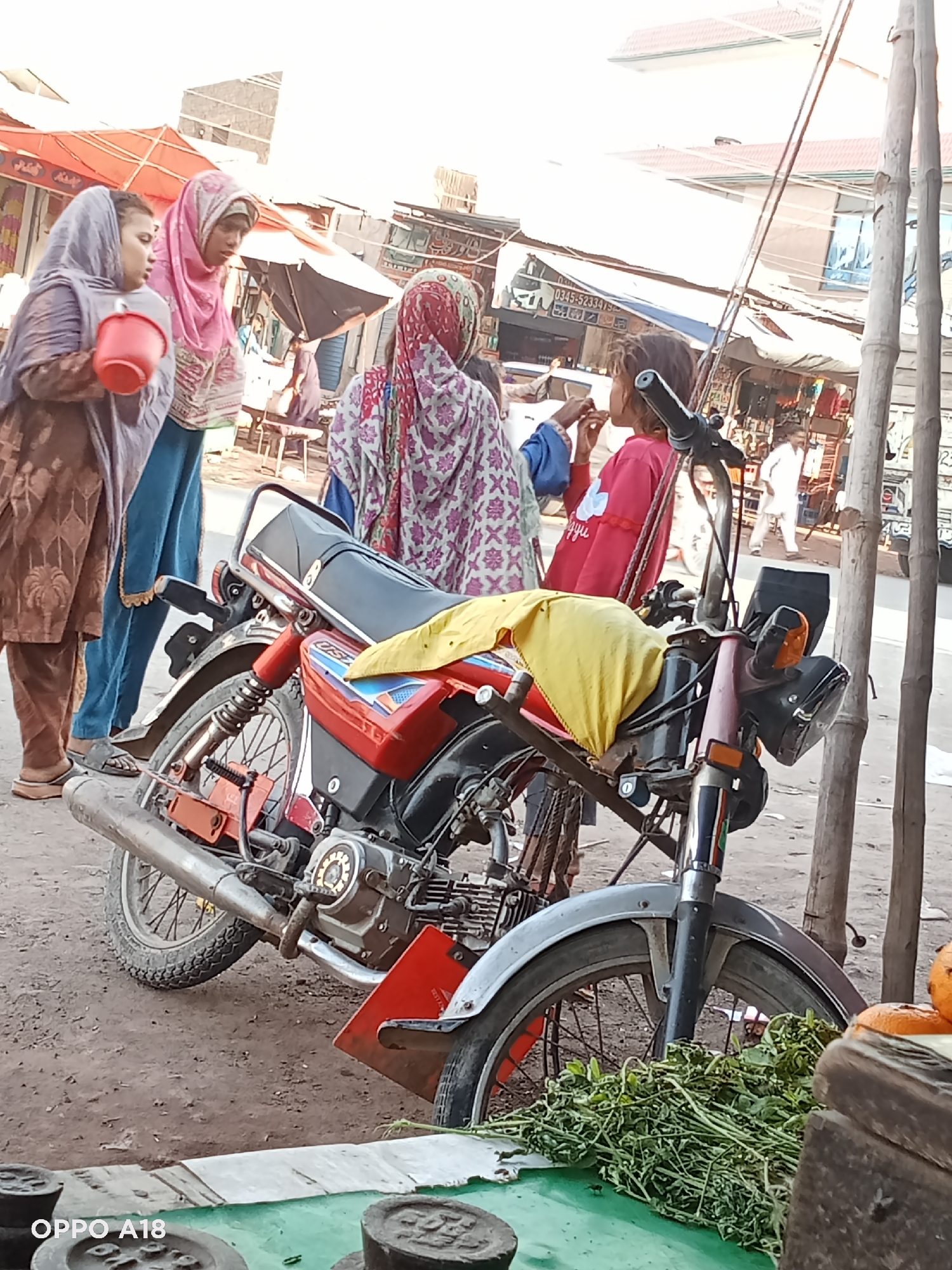 An outdoor market scene unfolds under bright daylight, featuring several individuals and market goods. In the foreground, a prominent red and black motorcycle is parked, leaning on its kickstand. A yellow cloth is draped over its handlebars, and green leafy vegetables are displayed on a stall surface directly in front of its front wheel. Alongside the vegetables, a few oranges are visible, and dark, round weights lie on the stall.
To the left, two women are engaged in conversation. One woman, wearing a brown patterned outfit and a light purple head covering, holds a red plastic bucket. Beside her, another woman in a pink head covering and a patterned top looks towards the first woman. Further to the right, with her back mostly to the camera, a third woman wearing a white, pink, and green patterned head covering and top stands near a young girl. The girl, dressed in a red top, appears to be eating something, holding her hand to her mouth.
The background reveals a bustling market environment with various stalls and shops, some featuring awnings and bamboo supports. Signs with text, including a partially visible phone number "0345-5233415" and some unreadable text in what appears to be Urdu or Arabic script, adorn the shopfronts. A man in a white outfit is seen walking in the mid-ground. The ground is a dusty, uneven dirt surface. The overall atmosphere suggests an ordinary, busy day at a local market. "OPPO A18" is visible in the bottom left corner, indicating the device used for the photograph.