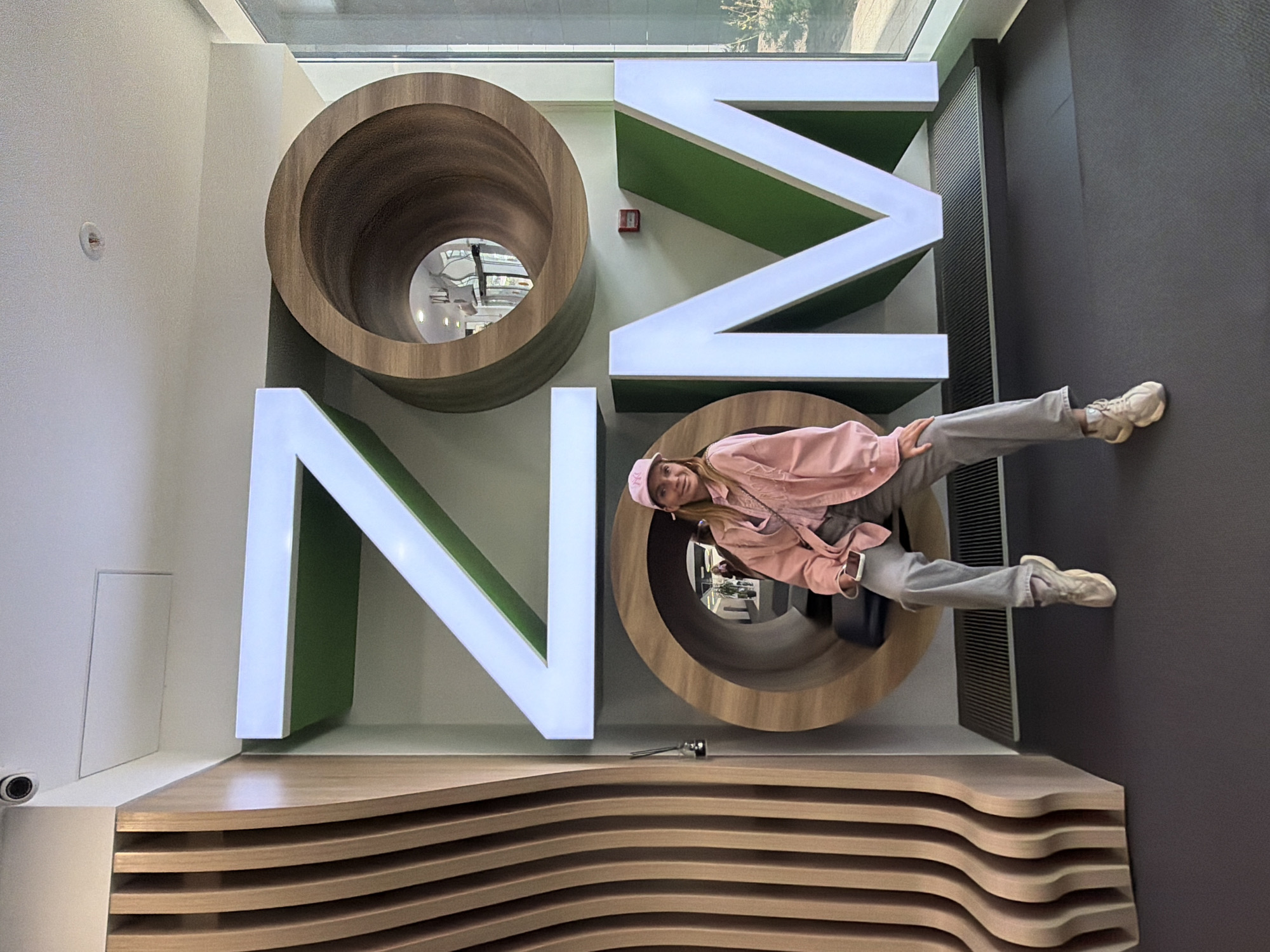 A horizontally oriented photograph captures a scene in what appears to be an interior setting with artistic installations, likely in Saint Petersburg, Russia, although specific city landmarks are not visible. The primary subjects are large three-dimensional letters forming a design, and a young woman who is partially positioned within one of the letters.
The backdrop includes elements such as a window, a gray wall, and a wooden installation that resembles layers or contours. The large letters are white with green interiors, and two of them are circular tubes with wooden texture. The young woman, wearing a pink jacket, gray pants, and sneakers, is oriented sideways, with her legs extending towards the gray wall.
The scene is well-lit, but lacks any distinctive details that would provide further information about the exact location or specific events.
