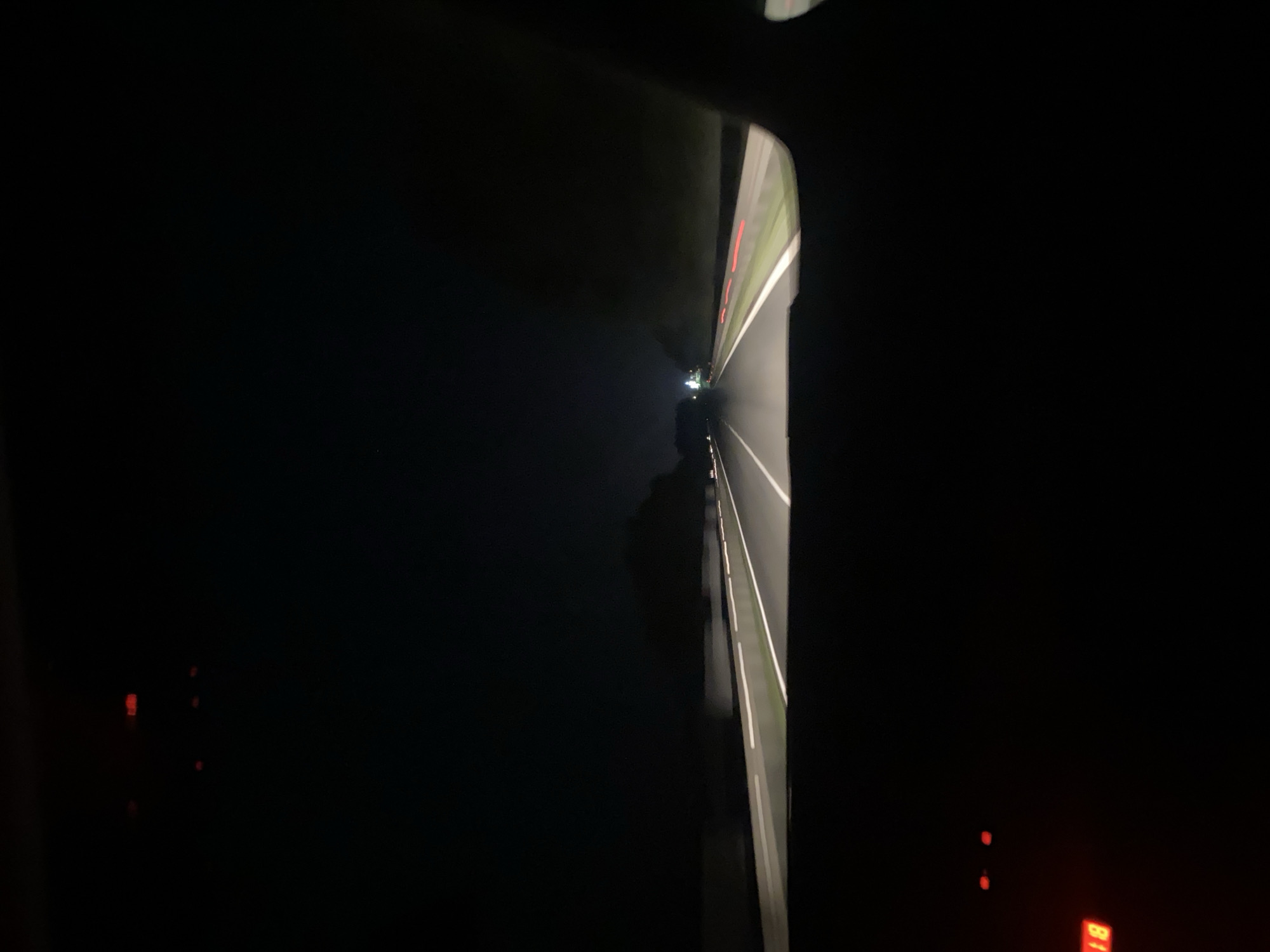 The image is a nighttime view from inside a vehicle driving on a highway. The highway has multiple lanes marked with white lines, and the road is illuminated by the vehicle's headlights. Distant streetlights and the taillights of other vehicles are visible. The surrounding environment is dark, with trees and foliage lining the sides of the road. In the foreground, parts of the vehicle's interior are visible, including a dashboard with some illuminated red indicators, one of which displays the number "18". The scene appears to be in Woskrzenice Duże, Poland, given the provided context.