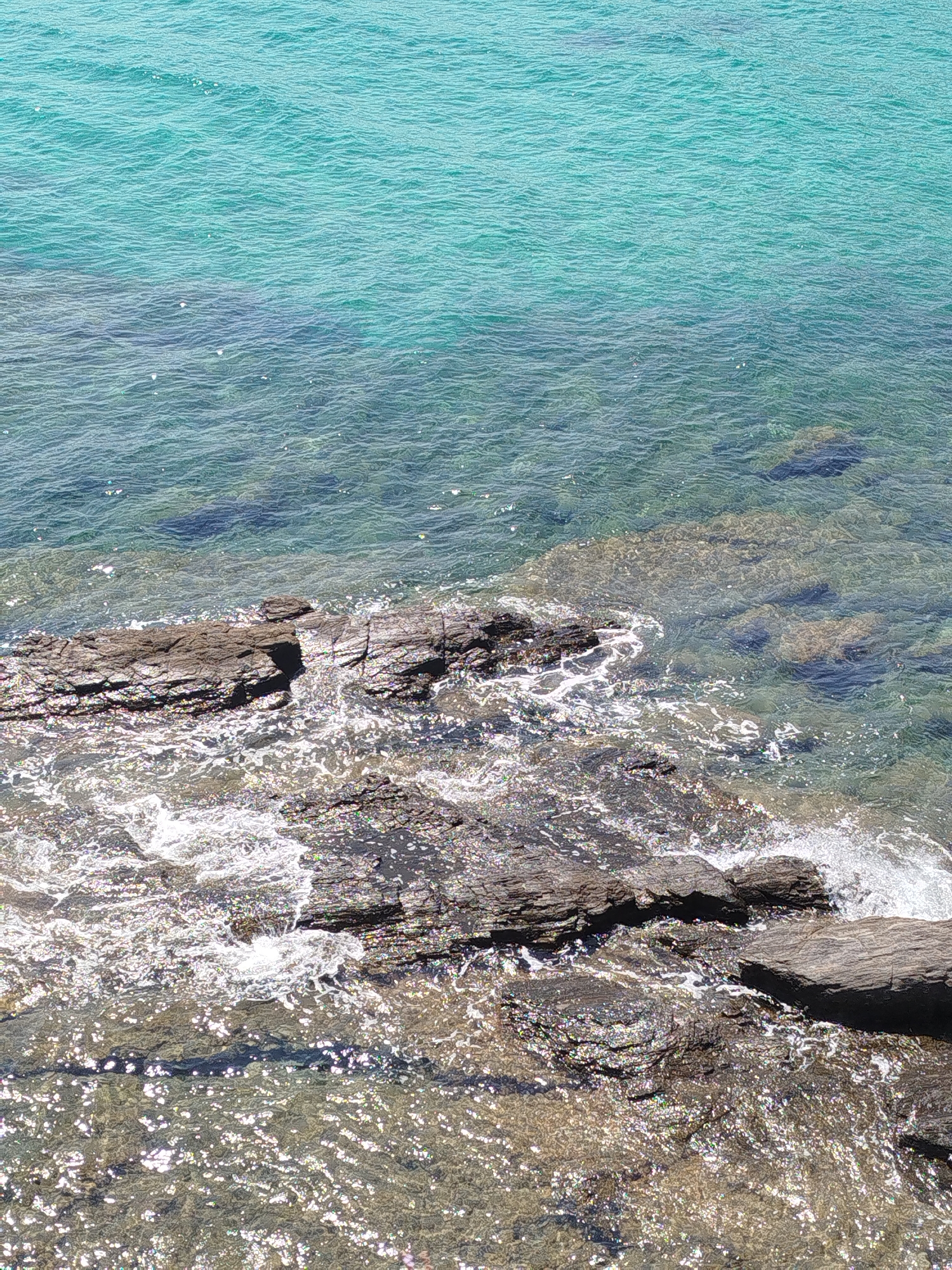 The image shows a rocky shoreline meeting clear, turquoise water under a sunny sky. The water is shallow near the shore, revealing submerged rocks and the sandy or pebbly seabed. Small waves are breaking against the dark, textured rocks, creating white foam and sparkling highlights on the water's surface. The deeper water exhibits a gradient of color from light turquoise to a deeper blue, with the seabed visible in patches. The scene captures a natural coastal environment. The location is within the Sithonia Municipal Unit, Greece, a region known for its scenic coastlines. There are no people or animals visible in the image.