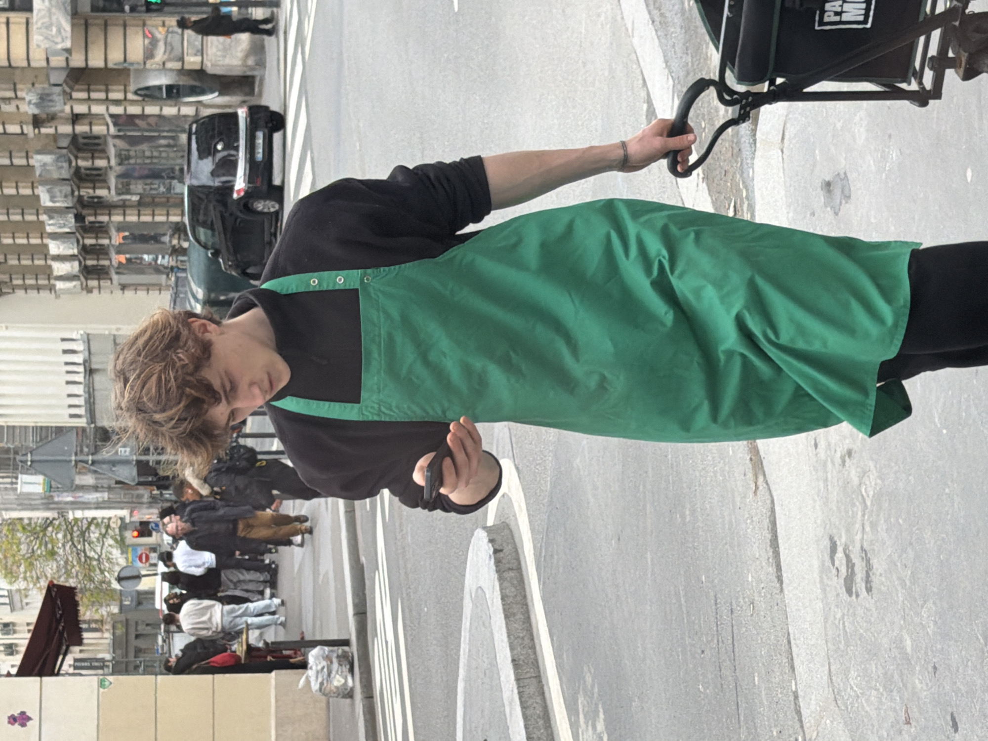 A young adult wearing a Starbucks apron is walking on a sidewalk in Paris, France. The individual is looking at a phone in their hand and holding the handle of a black cart. In the background, there are pedestrians walking in the opposite direction, a car on the street, and Parisian buildings. The environment appears to be a busy urban area during daylight.