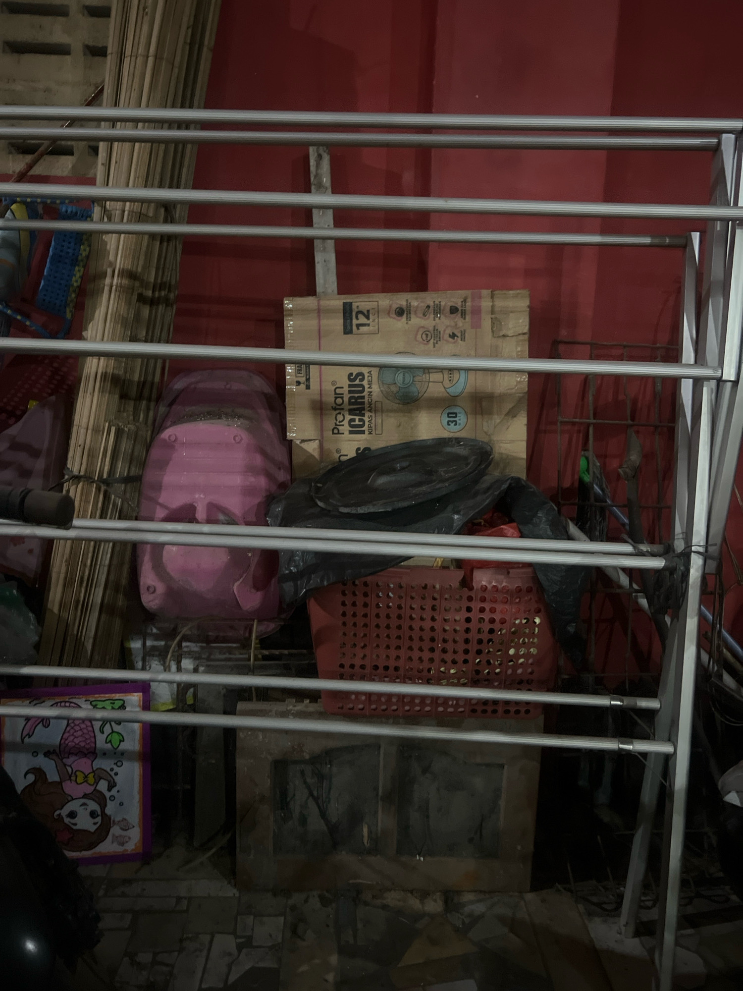 The image depicts an outdoor storage area or alleyway, likely behind a building in Tlogoadi, Indonesia. In the foreground, a silver metal drying rack or shelving unit obstructs the view of several stacked items. Behind the rack, a stack of light brown cardboard boxes is visible, with one box prominently displaying "Profan ICARUS Kipas Angin Meja 12 INCH" and a fan graphic. To the left of the boxes, a large, pink plastic object, possibly a laundry basket or storage container, is partially visible. Below this, a red plastic crate filled with items is seen. Further left, a framed item with a drawing of a mermaid is propped against the wall. The background is dominated by a red wall and a collection of vertical wooden or bamboo poles. The ground appears to be paved with rough stones or concrete. The overall scene suggests a disorganized but functional storage space.