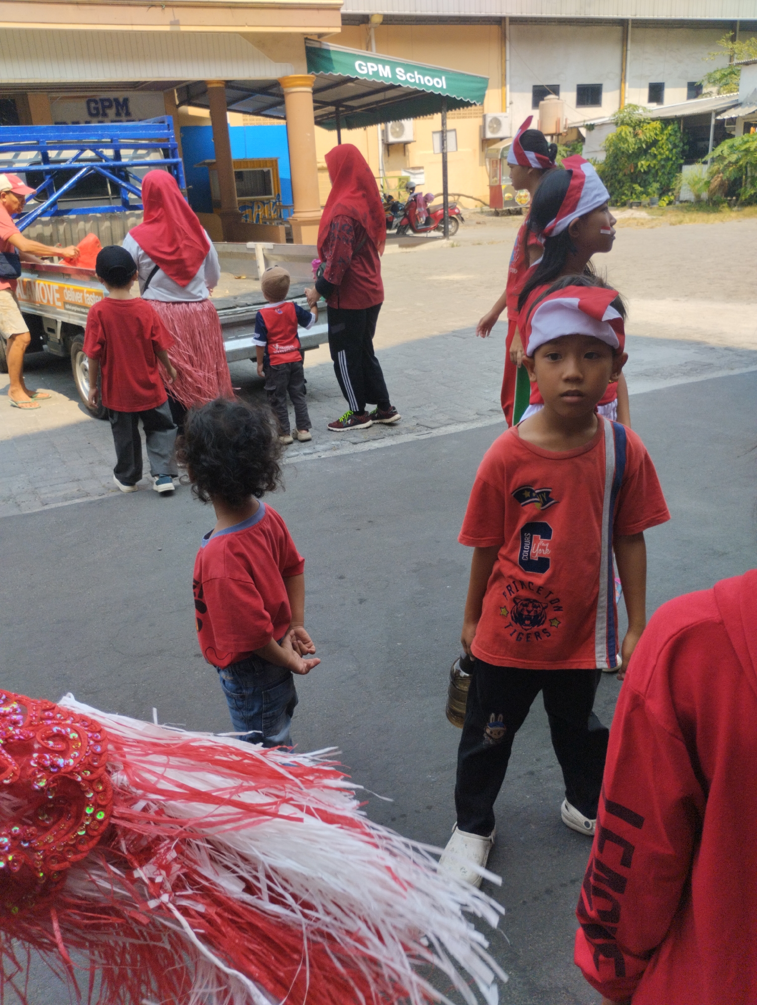 In front of the "GPM School" building in Bekasi, Indonesia, a group of people, primarily children, are gathered. A red and white decorative fringe with sequins is prominent in the foreground. Several children are wearing red shirts and some have red and white headbands, suggesting a festive or school event. One boy in the foreground, wearing a red shirt with graphics that include "Princeton Tigers," is holding a bottle. Another child with curly hair is standing behind him, facing away from the camera. In the background, adults in red headscarves and children are near a truck. Red motorcycles are parked near the school entrance. The ground is paved, and the environment appears to be an outdoor school or public space.