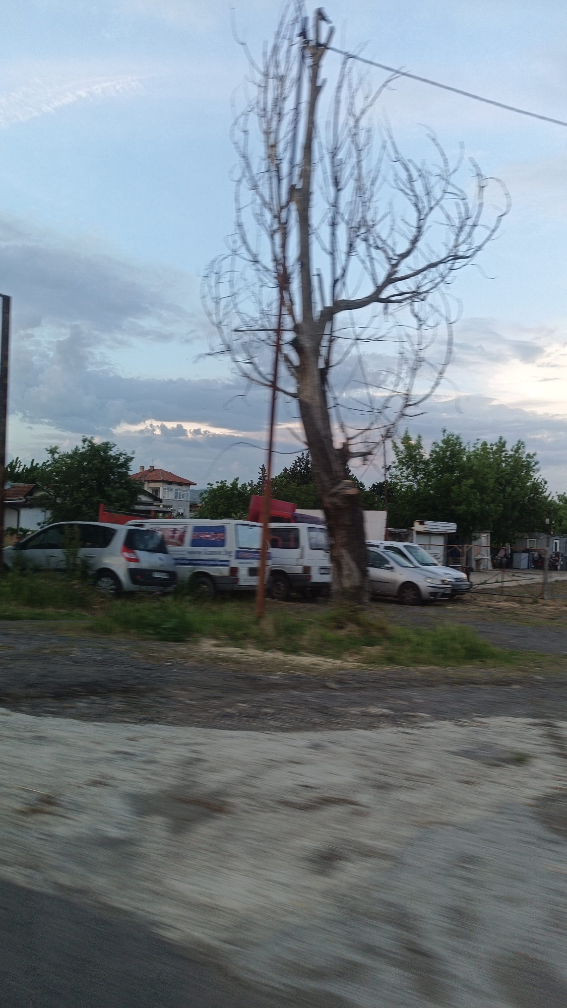 The image shows a roadside scene. In the background are several parked vehicles, including vans and cars. Behind the vehicles are green trees and a building with a red roof. In the foreground, there's a blurry patch of asphalt or concrete. A tall, bare tree stands near the roadside. The sky is cloudy with patches of blue. The location may be in Burgas, Bulgaria, as specified in the request. There are people gathered near some of the parked vehicles.