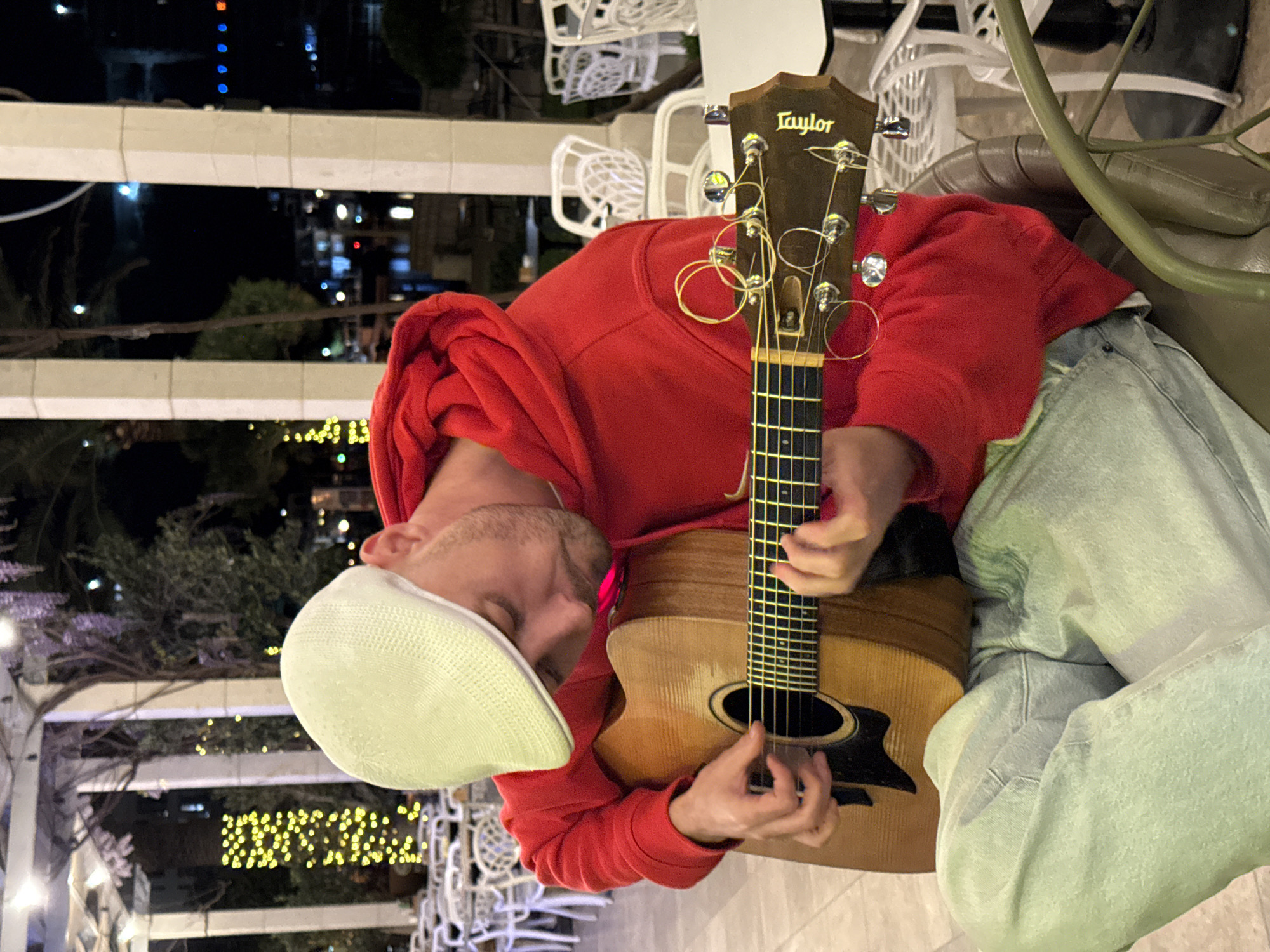 A man in a red hoodie and a white flat cap plays a Taylor acoustic guitar in an outdoor setting. The man sits with light blue jeans on, his head close to the guitar's neck, and his hands strum the strings. The guitar's headstock is visible, displaying the "Taylor" logo. The scene is at night, with city lights visible in the background. The location is Rafailovići, Montenegro. White chairs and tables, along with a decorative pergola structure, surround the man.