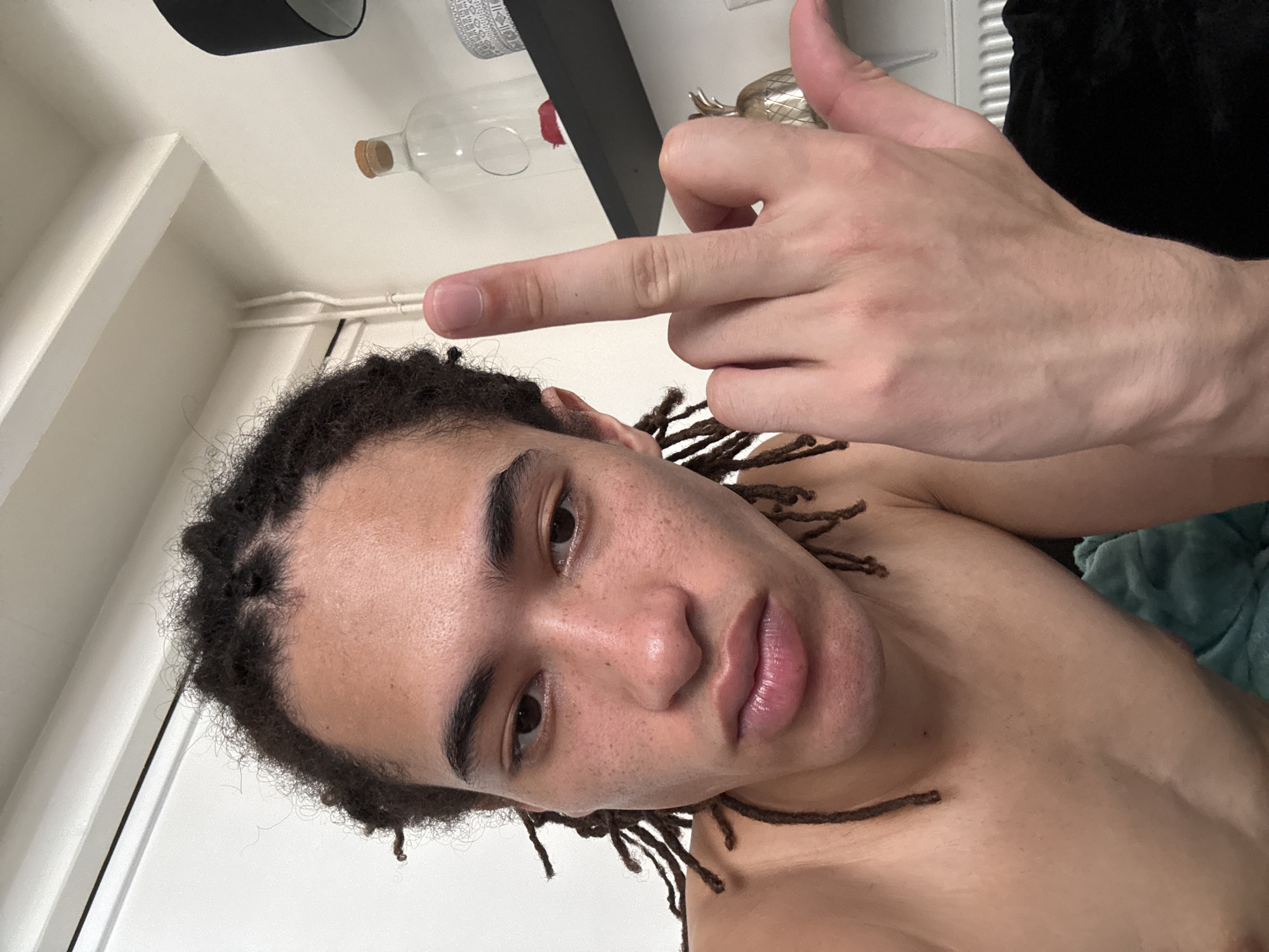 The image shows a close-up, selfie-style shot of a young person with dreadlocks, shirtless and looking at the camera with a neutral expression. They are holding up their middle finger. The background appears to be an indoor setting, possibly a bedroom or living area, with a white wall, a shelf with a bottle and decorative items, and a radiator visible. There are no discernible landmarks or elements that would indicate the location as Paris, France.