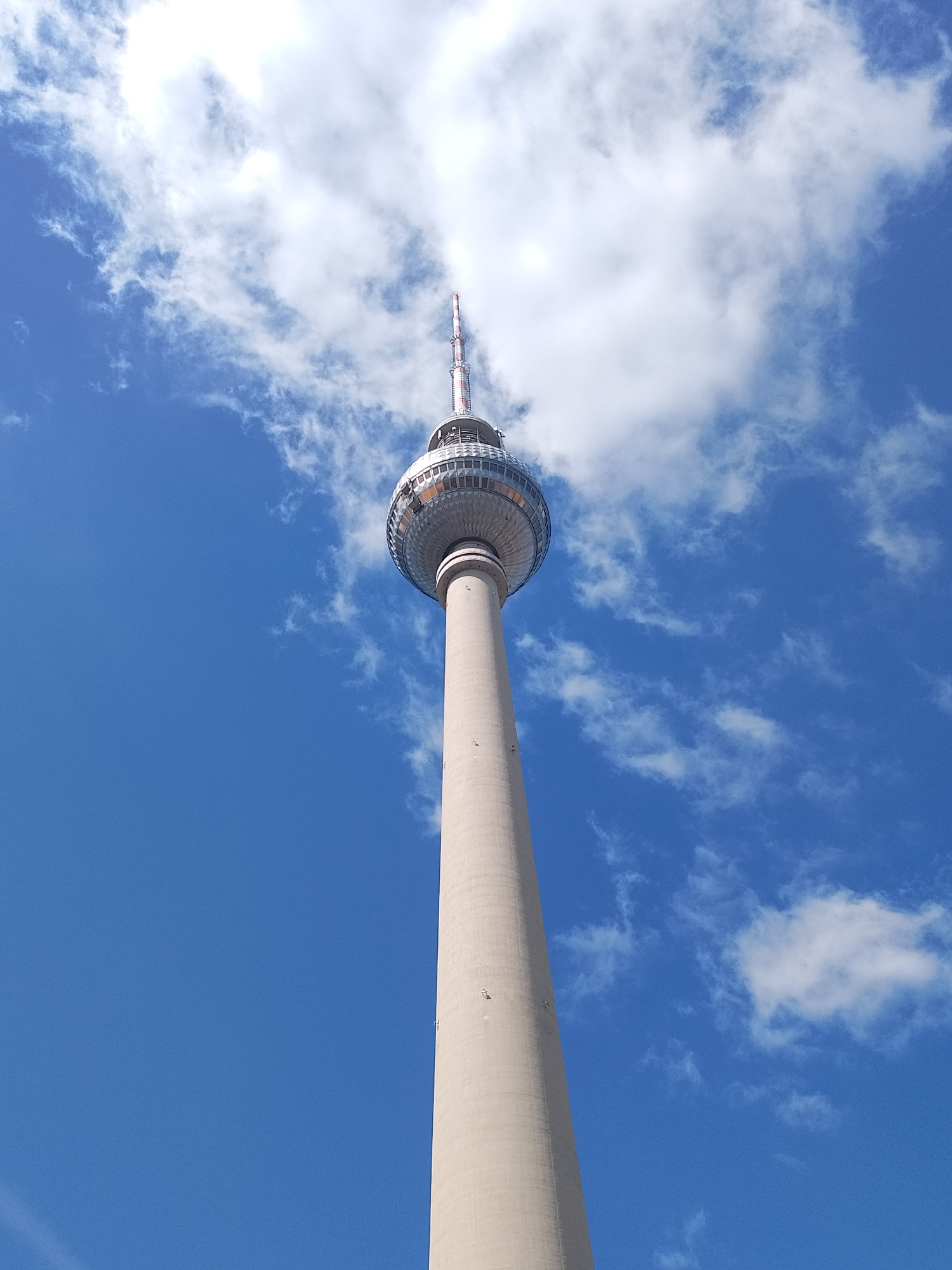 The image shows the Berlin TV Tower in Berlin, Germany. The tower's tall, cylindrical base leads to a spherical observation deck and a red-and-white antenna. The sky is blue with scattered white clouds.