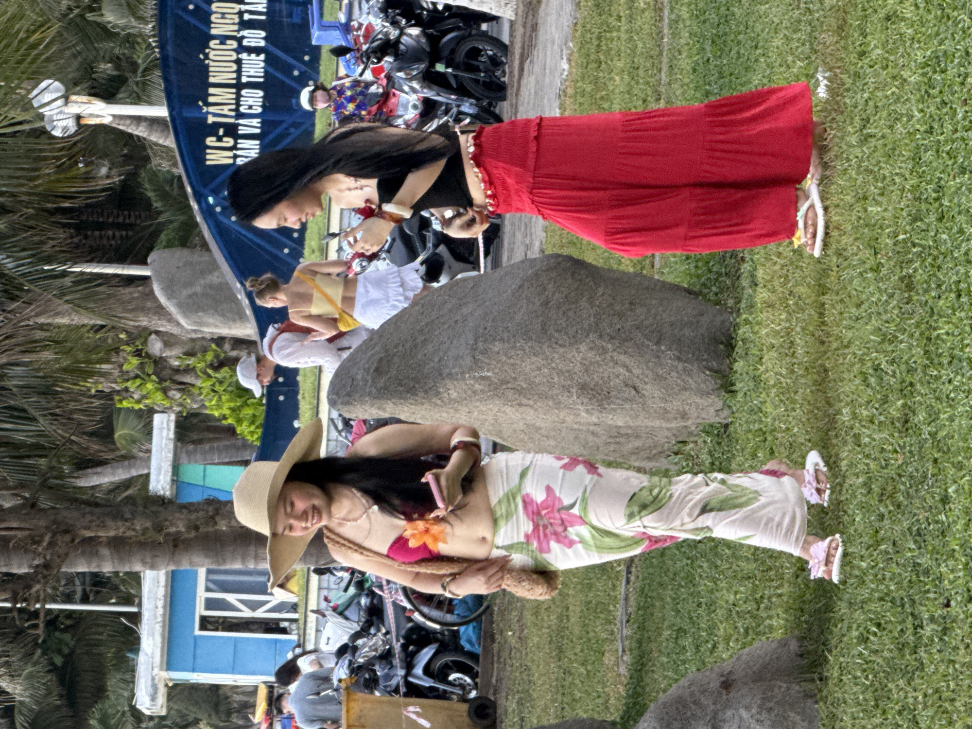 In Nha Trang Ward, Vietnam, two women are posing outdoors near a blue structure with Vietnamese text, "WC - TẮM NƯỚC NGOÀI VÀ CHO THUÊ ĐỒ TẮM". The woman in the foreground, wearing a wide-brimmed straw hat, a pink bikini top, and a floral print skirt, smiles widely. She has a flower on her bikini top and is holding a woven bag. She is standing next to a large rock. Behind her, another woman in a black top and a red maxi skirt is also posing, her hand resting on her stomach. Several motorcycles are visible in the background, suggesting a public area. The scene appears to be outdoors, with a grassy area and palm trees. The lighting suggests it is daytime. The overall mood is relaxed and summery.