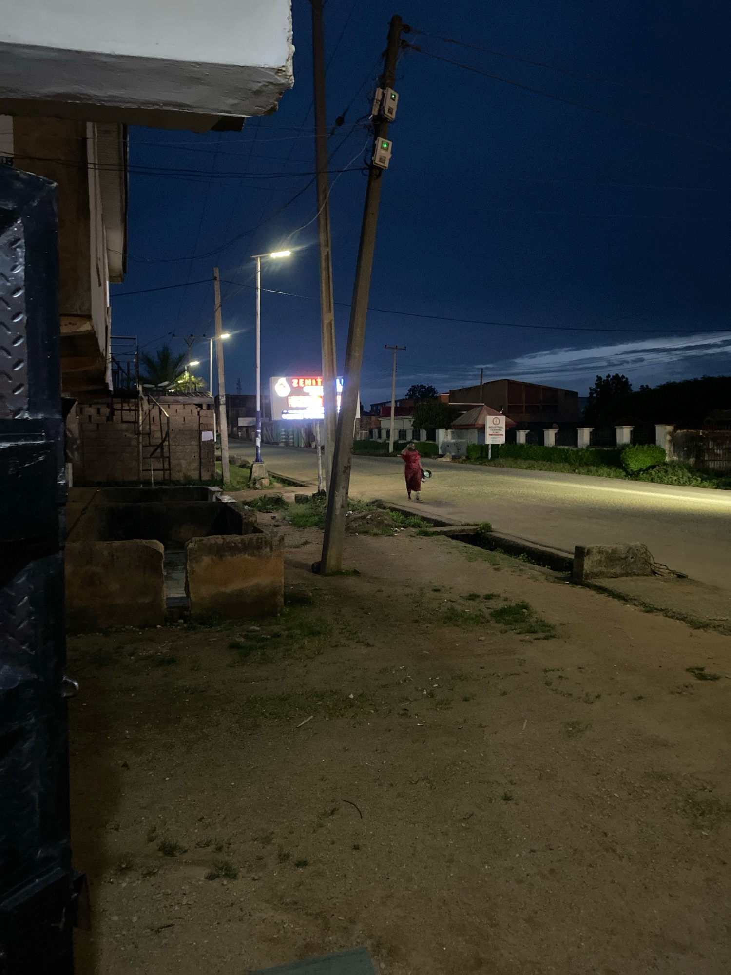 The image depicts a street scene at dusk in Jos, Nigeria. A lone figure wearing a red garment is walking along the side of a paved road, which is illuminated by streetlights. The figure appears to be carrying something in their left hand. On the left side of the image, a building with a prominent overhang is visible, with a dark, textured metal structure in the foreground. Several utility poles with electrical equipment are present along the roadside. In the background, there are buildings and vegetation. The sky is a deep blue, with some wispy clouds. The overall impression is that of a quiet urban street at the end of the day.