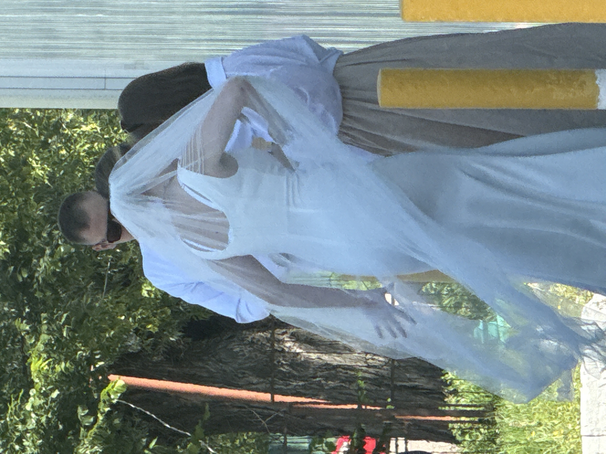 The image shows three individuals outdoors, with a focus on a person in a white wedding dress and veil. The wedding dress is sleeveless with a low back and a flowing veil. A man wearing a white long-sleeved shirt and sunglasses stands behind the bride, his arm around her. To the right of the bride, another person, partially obscured, wears a white long-sleeved shirt and a light brown skirt.
The background consists of lush green trees and foliage. In the foreground, there are two yellow posts with white bases. The overall setting appears to be a public area with vegetation, possibly near a building with a vertical ribbed texture on the right side. No specific landmarks of Yakhroma, Russia, are identifiable in this image. The lighting suggests it is daytime. The individuals appear to be in a procession or posing for a photograph.