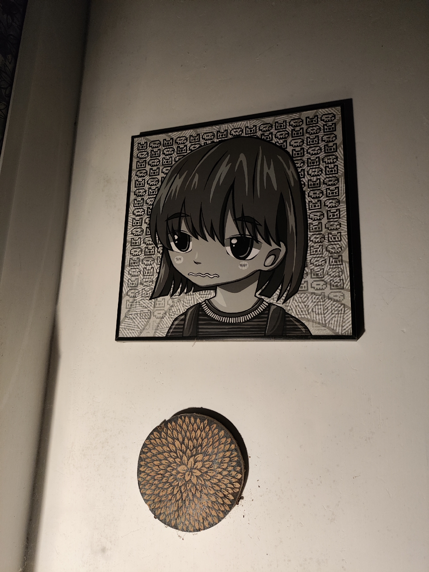 A framed black and white anime-style drawing of a young girl with large eyes and a worried expression hangs on a textured, off-white wall. The drawing is set against a background filled with repeating pixelated patterns. Below the drawing, a round, dark wooden coaster with a carved floral design is affixed to the wall. The scene is illuminated by artificial light, casting shadows on the wall to the left. This image was taken in the city of Kepung, Indonesia.