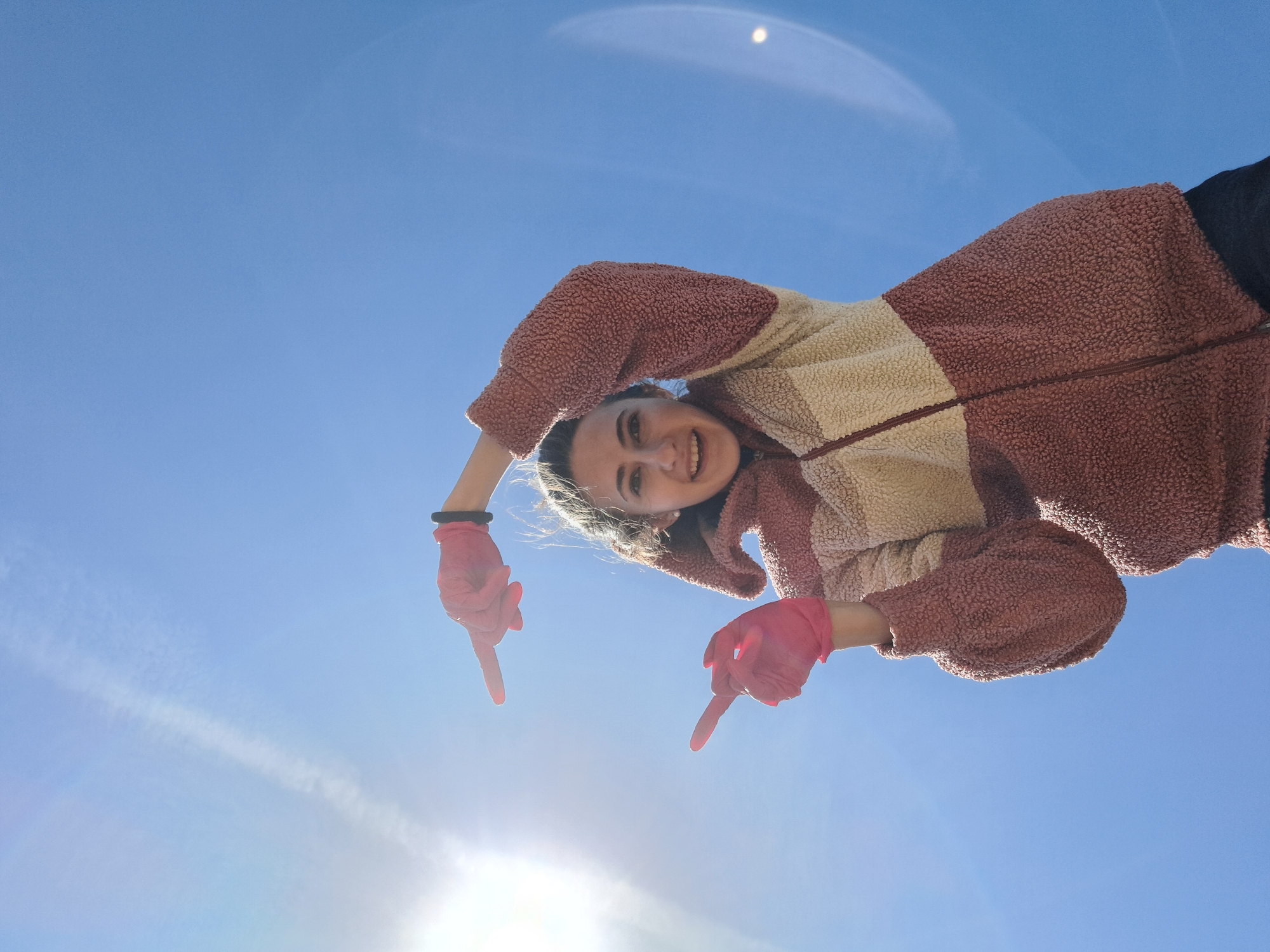 A young woman is posing against a clear blue sky. She is wearing a brown and beige fleece jacket and pink gloves. Her arms are raised, and she is pointing downwards with both index fingers. A bright light source, possibly the sun, is visible in the lower part of the frame, causing a lens flare. The location is indeterminate.