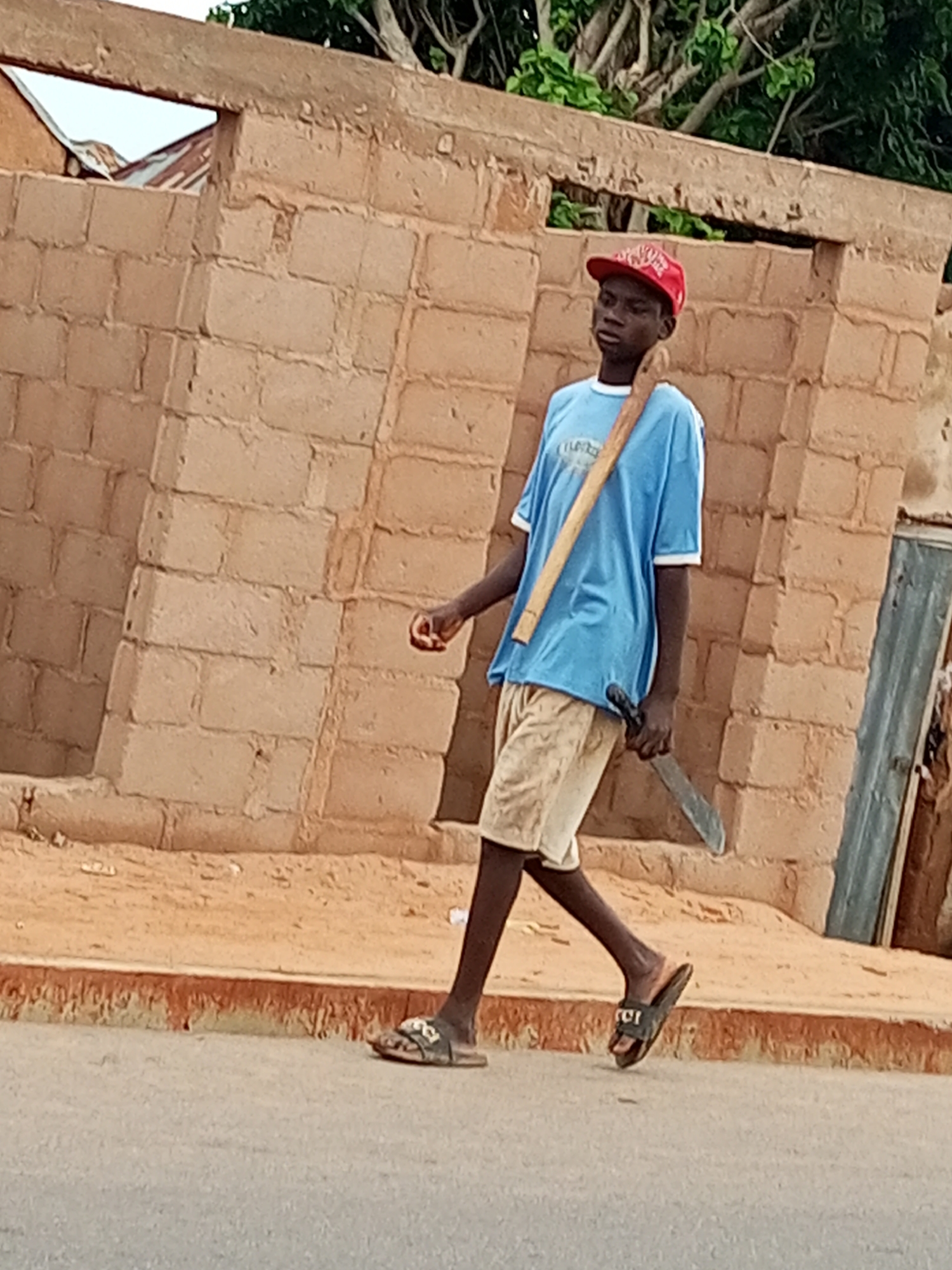 A young male wearing a blue t-shirt, light-colored shorts, and a red baseball cap walks on a paved surface in front of a light brown brick wall. A wooden tool is slung over his right shoulder, and he is carrying a machete in his right hand. The background shows some vegetation and part of a building. The scene appears to be in an outdoor urban environment, possibly in Gombe, Nigeria.