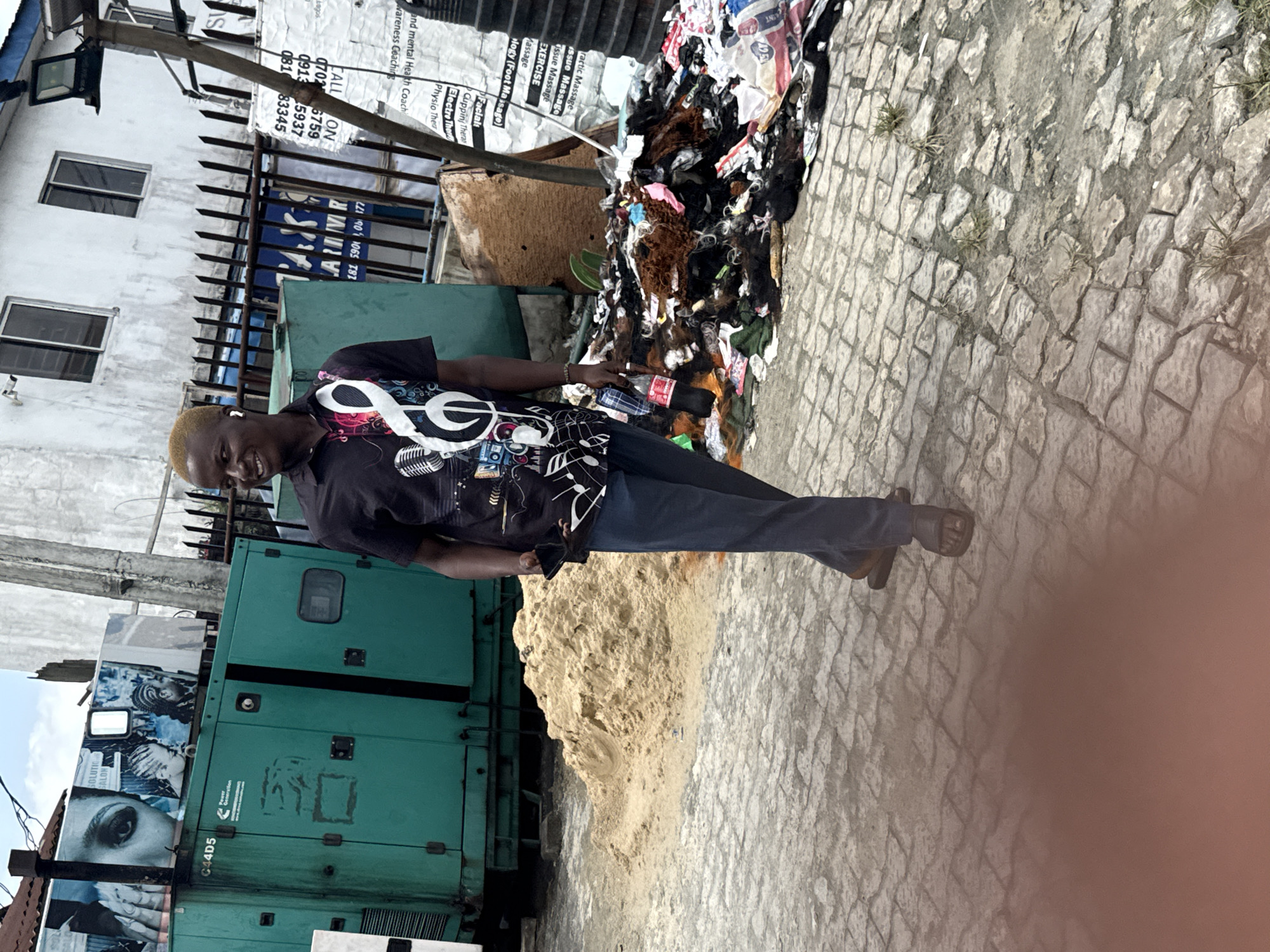 A man with blond hair is standing on a paved street next to a pile of debris, near a green generator in Ado, Nigeria. The man wears a t-shirt with musical note designs and dark-colored pants. The pile of debris includes dark-colored hair extensions. Behind the man, on a building wall, are posters with text: "Practic Massage", "Exercise", "Facial", "Physio Ther", and contact information. The generator has the text "C44D5" and a salon advertisement with an image of a woman's eye. A pile of sand is next to the generator.