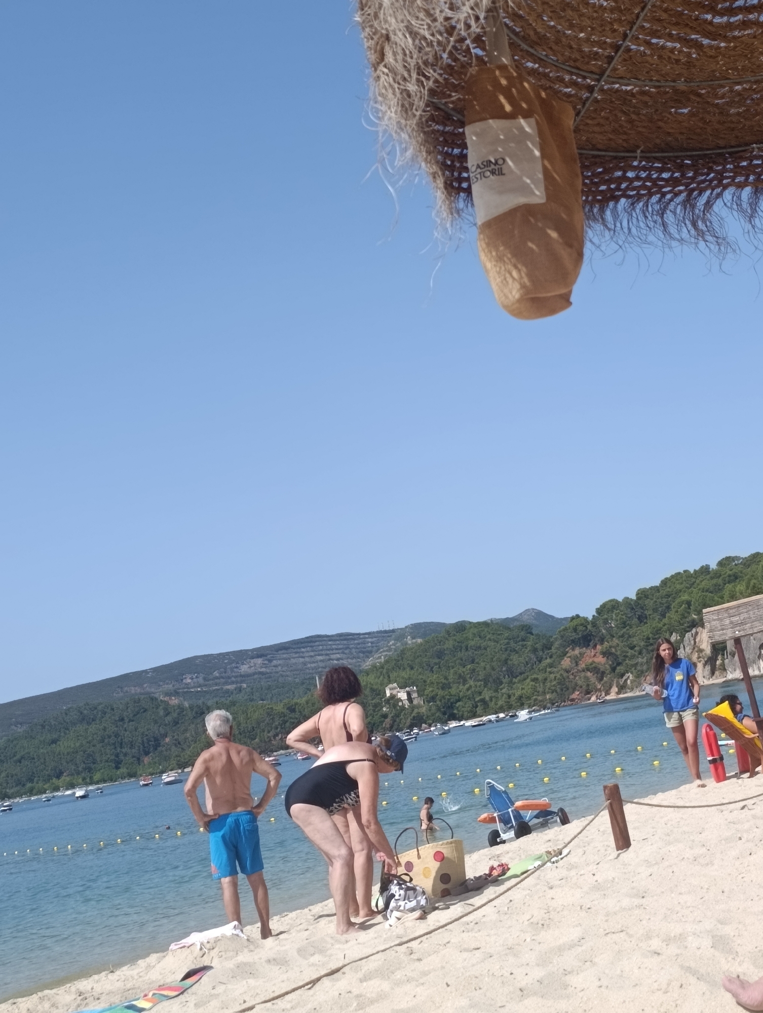 The image captures a sunny day at a beach in Setúbal, Portugal, likely along the Sado Estuary. In the foreground, the sandy beach is dotted with beachgoers. An older man with gray hair stands facing the water, wearing blue swim trunks. Beside him, a woman in a black bikini is bent over, interacting with a bag. Further back, a young woman in a blue t-shirt and shorts stands near a lifeguard station, holding a bottle of water.
The ocean stretches out behind the beach, dotted with numerous small boats. The coastline is lined with lush green hills, and the sky is a clear, bright blue. A portion of a straw umbrella and a decorative element with the text "CASINO ESTORIL" are visible hanging from above, suggesting a proximity to a beach club or establishment. The scene depicts a typical summer day at a coastal resort, with people enjoying the water and the sunshine.