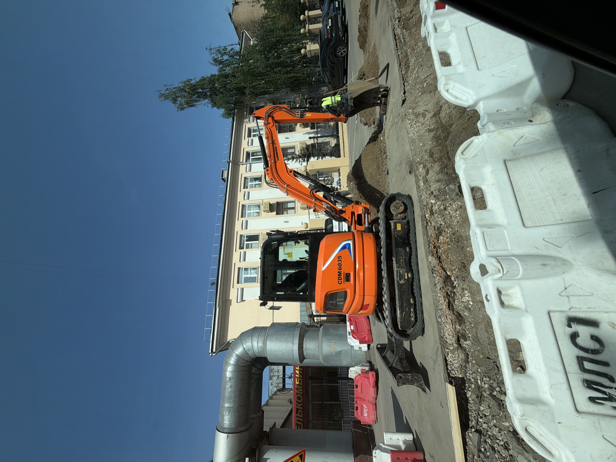 The image shows a construction site in Moscow, Russia, featuring an orange mini excavator digging a trench. The excavator is positioned on a paved surface next to a large, silver, curved pipe. A construction worker wearing a yellow vest is visible near the excavator's bucket. White and red plastic barriers surround the work area. A building with a light-colored facade is in the background. A store with a sign including the word "ЕЛЬКОМБИ" is partially visible behind the pipe. A street with parked vehicles runs parallel to the construction. The sky is clear and blue.
