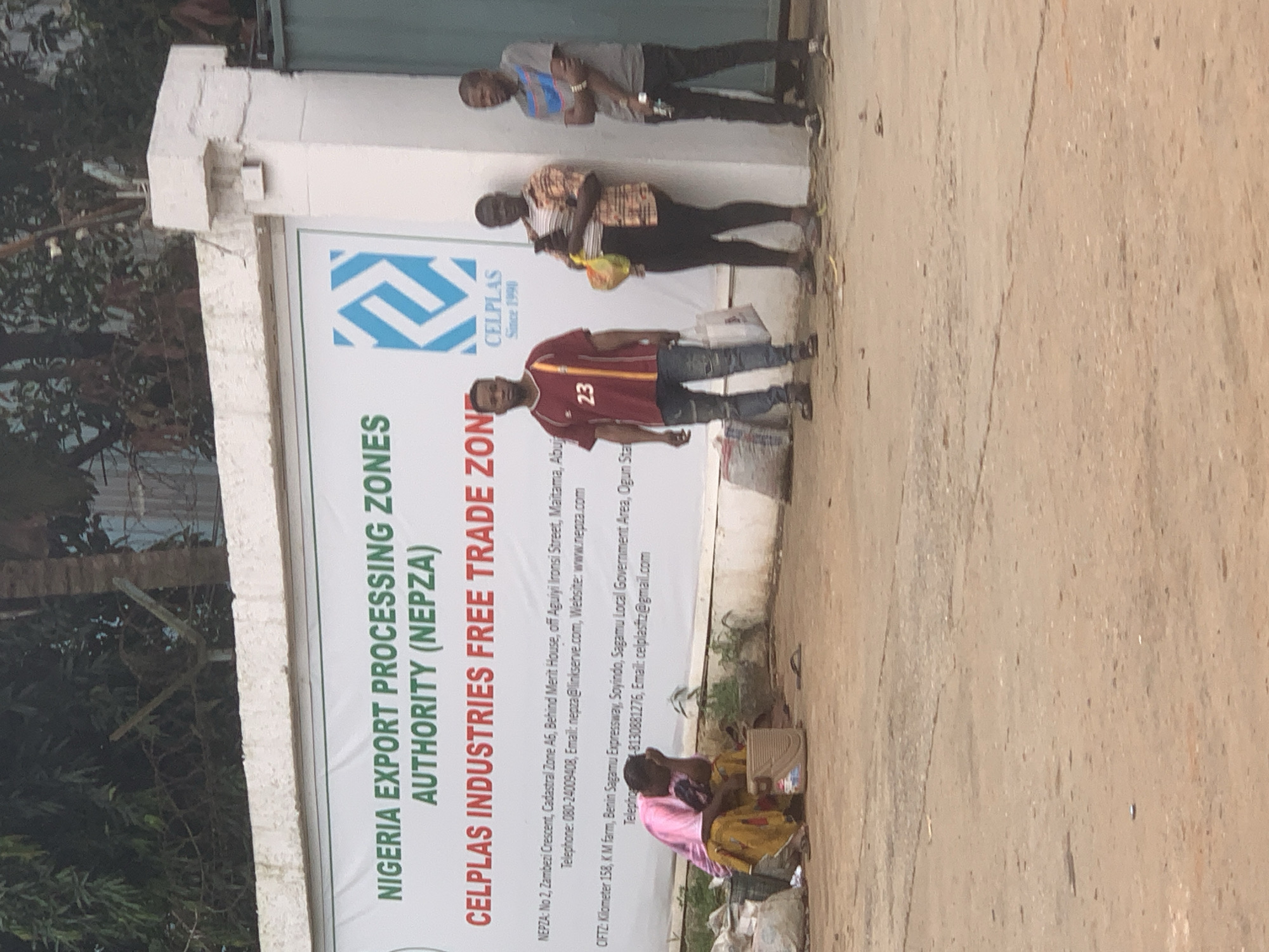 The image depicts the entrance to a free trade zone in Shagamu, Nigeria. A large banner is prominently displayed, featuring the text "NIGERIA EXPORT PROCESSING ZONES AUTHORITY (NEPZA)" and "CELPLAS INDUSTRIES FREE TRADE ZONE". Below this, contact information for NEPZA and CIFTZ (export processing zone) is visible, including addresses, phone numbers, and email addresses.
In the foreground, there are several people. On the left, a woman in a pink top and patterned skirt is seated, appearing to be resting or waiting. Next to her are some bags and a plastic container. To her right, three men are standing. The man on the far left is wearing a maroon shirt with the number "23" on it, blue jeans, and carrying a white bag. The middle man is wearing a patterned shirt and dark pants, holding a yellow bag and a mobile phone. The man on the far right is wearing a grey t-shirt with a blue stripe and dark pants.
The environment appears to be an outdoor setting with a concrete ground. The background shows trees and a white building structure associated with the free trade zone entrance. The overall impression is a functional industrial zone entrance with people present, possibly waiting for entry or interacting.