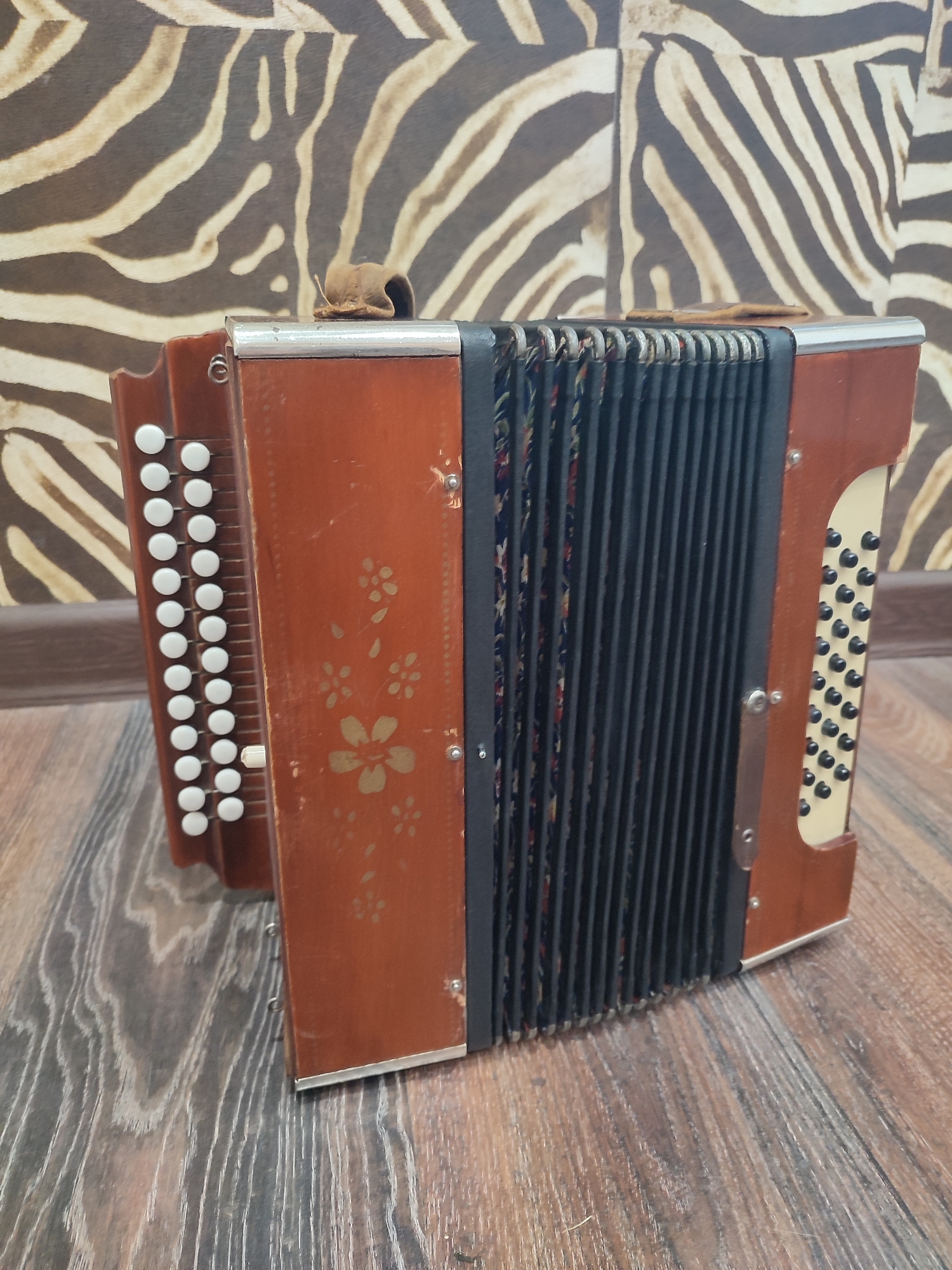 An accordion, primarily brown with black bellows, sits on a wooden floor. The accordion has white buttons on the left side and black buttons on the right side. Decorative floral patterns are visible on the brown casing. In the background, a wall displays a pattern resembling zebra stripes. The scene is indoors.