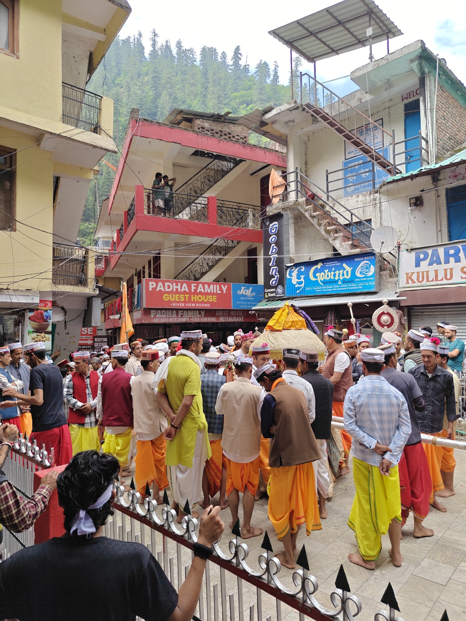 The image depicts a bustling street scene in Kasol, India, centered around a large gathering or procession of men. The majority of the participants are dressed in traditional Himachali attire, including saffron, yellow, and white dhoti-like lower garments (locally known as gachhas) and distinctive round Kullu caps (topis) adorned with colorful bands. Some men wear vests or shirts over their traditional drapes, and several are barefoot, emphasizing the traditional nature of the event.
At the heart of the crowd is a decorated palanquin-like structure, richly adorned with yellow and purple fabric, suggesting it carries a deity or sacred object. This indicates a religious procession, common in the local festivals of the region.
The setting is a narrow, enclosed street lined with multi-story buildings, painted in various colors such as yellow, red, and off-white. Balconies on these buildings host onlookers observing the activity below. In the background, lush green mountains covered with dense pine forests rise, characteristic of the Himalayan foothills. Numerous electrical and communication wires crisscross overhead. The scene unfolds during the daytime under an overcast sky, providing soft, diffused lighting.
Visible text on the buildings identifies various businesses. Prominent signs include "PADHA FAMILY GUEST HOUSE," advertising "RIVER VIEW ROOMS," "HOT BATH WITH SHOWER," and "RESTAURANT." Another large blue sign reads "Gobind Garments & Shawls," specifying "100% PURE WOOL" products like "Handloom Kullu Shawls, Stoles, Jackets, Cardigan, Ladies Top, Kurti, T-Shirts, Track Pants, Kids Garments & Cosmetics Etc." with a phone number "98825 85555." Other partial signs include "Jal Joy of India," "PAR KULLU S," and "WELCOME." In the foreground, a metal railing separates the viewer from the procession.