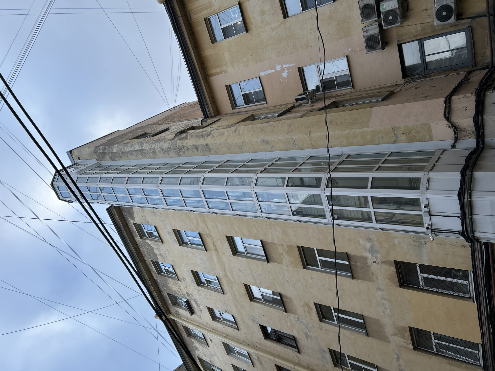 The image captures an upward-angled view of urban buildings in Saint Petersburg, Russia, under a bright, overcast sky. The scene is dominated by two multi-story buildings with weathered facades.
The building on the left exhibits a pale yellow-tan facade with numerous rectangular windows, many featuring white frames and reflecting the light sky. A prominent, long, and multi-tiered glass and white-framed enclosure, resembling an extended balcony or an enclosed walkway, projects diagonally upwards from a corner, narrowing as it ascends between the two structures.
The adjacent building, also displaying a similar faded yellow-tan hue, shows visible signs of wear, including patches of peeling paint and general discoloration. This facade features several windows of varying sizes, some with dark frames, and a cluster of outdoor air conditioning units mounted on its upper section. Dark electrical cables and conduits run vertically along its surface, often bundled together.
Numerous thin black utility wires crisscross the pale blue, somewhat cloudy sky overhead, connecting unseen points and contributing to the dense urban atmosphere. The perspective is from below, looking up towards the upper stories and the sky. The lighting suggests a daytime scene, likely an overcast day, providing soft and diffused light. No people or specific activities are depicted. There is no visible text within the frame. The overall impression is of a lived-in, utilitarian urban environment.