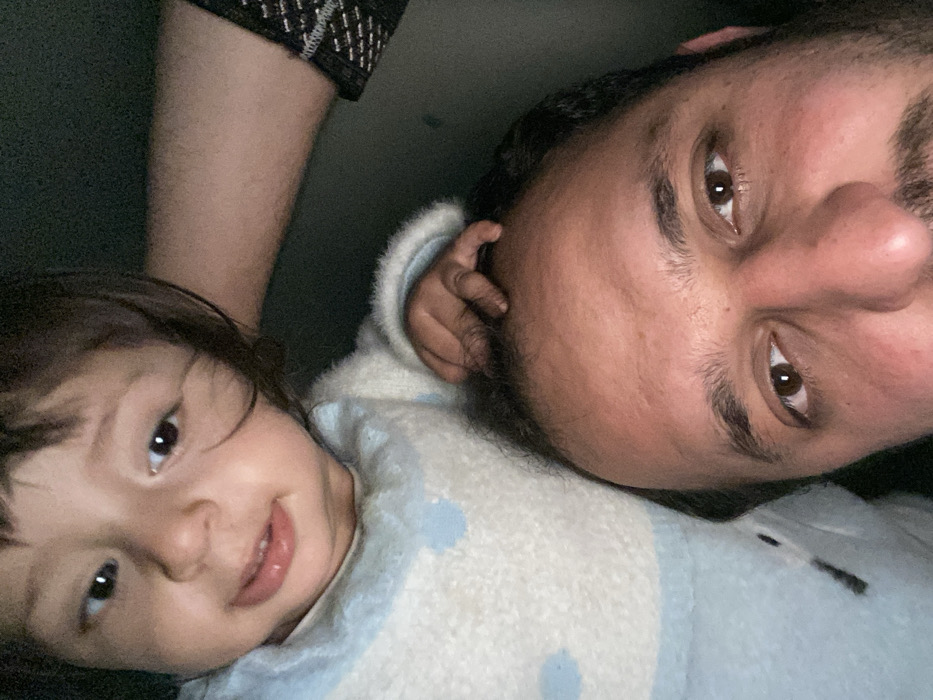 A close-up selfie captures a man and a child. The man, with dark hair and a beard, looks directly at the camera. The child, with dark hair and a slight smile, is positioned next to him. They are both lying down, possibly on a bed or blanket. The child is wearing a light blue and white patterned sweater. The man's arm, covered in a patterned sleeve, is visible behind the child. The overall image is slightly dark.